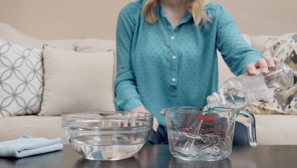 Woman pouring liquid cleaning solution into measuring cup. Glass bowl sitting beside measuring cup on a table.
