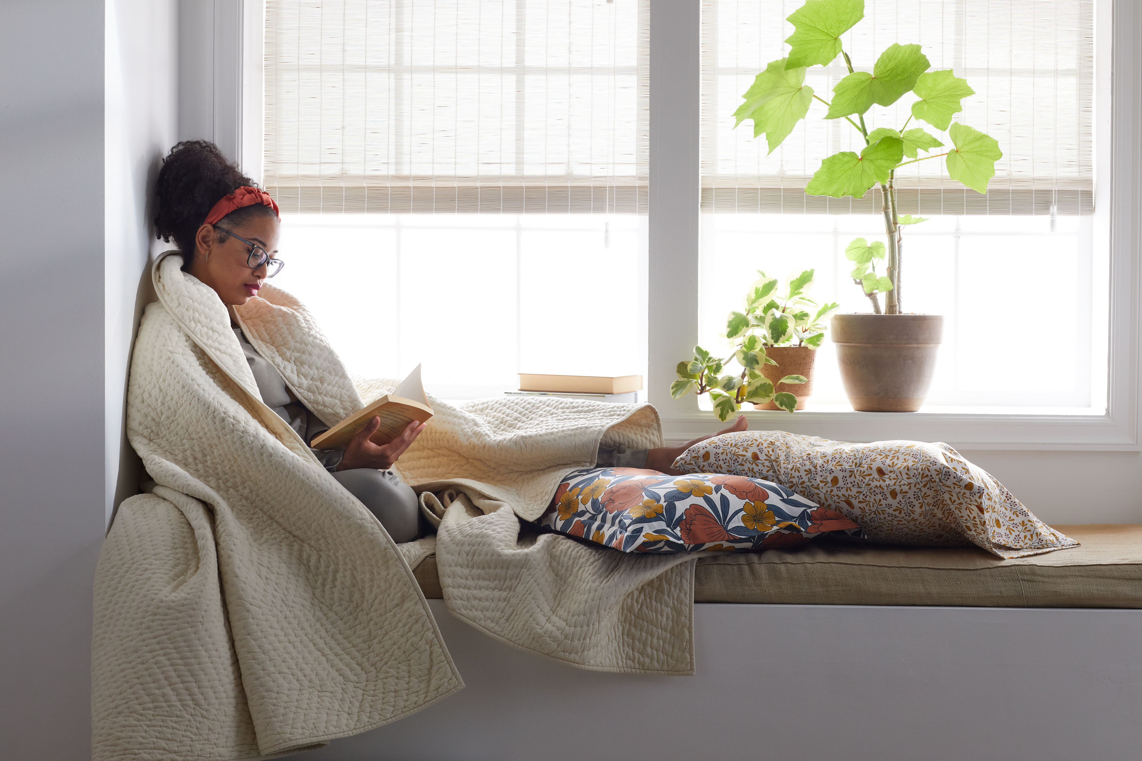 a woman reading a book in a cozy nook. woven wood shade window treatments and two houseplants are behind her