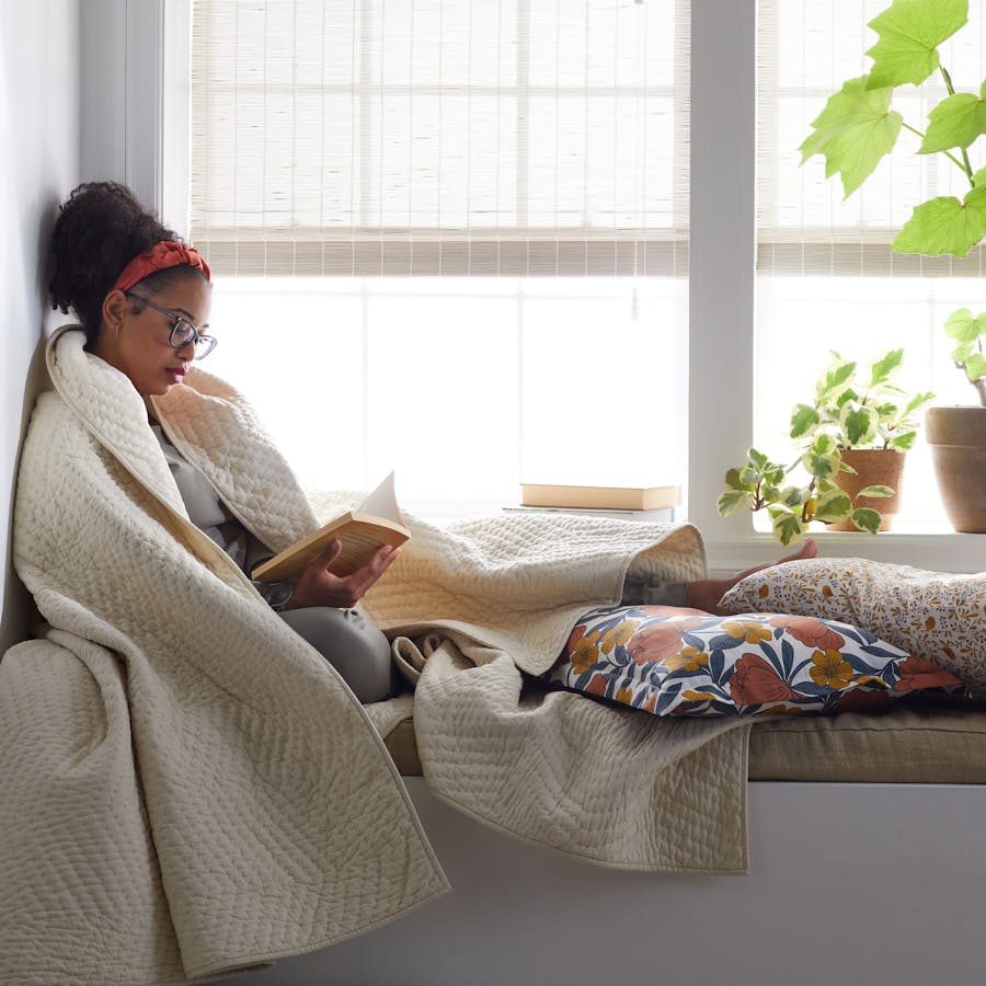 a woman reading a book in a cozy nook. woven wood shade window treatments and two houseplants are behind her