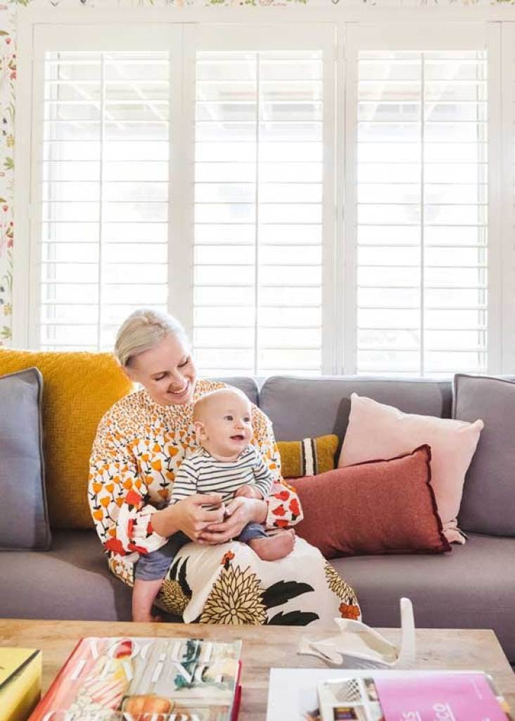 Woman holding baby boy in colorful living room in front of large window with white plantation shutters