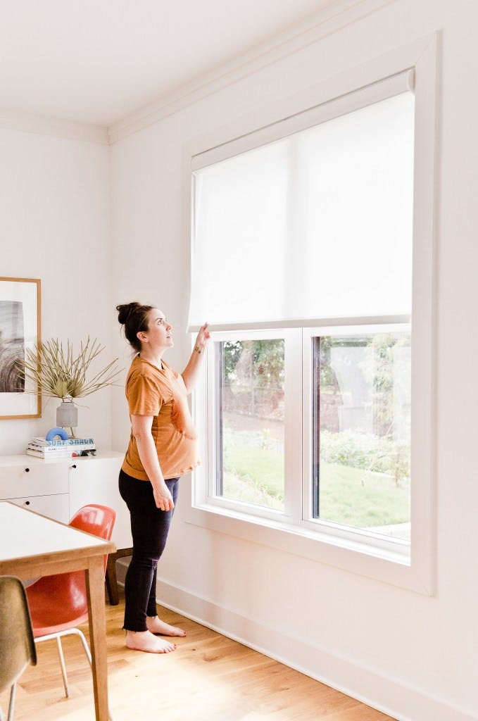Pregnant woman opens cordless white roller shades on large window