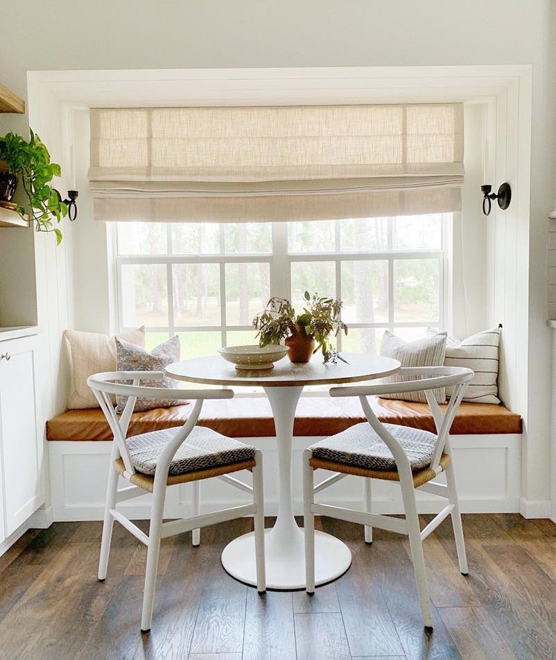 Nook dining area with cushioned bench seat and chairs. Decorated with houseplants and a beige roman shade.
