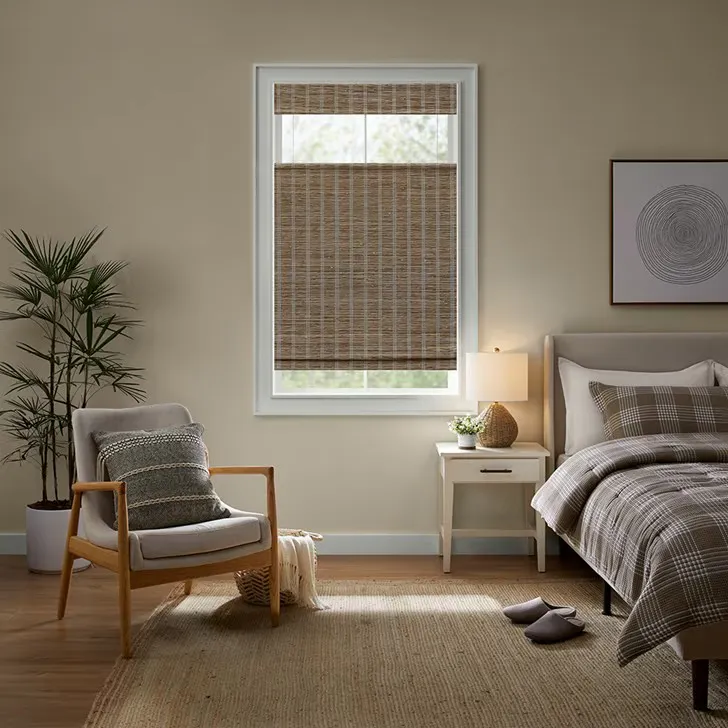 Blinds.com Woven Wood Shades in a natural woven texture, partially raised in a cozy, neutral bedroom with warm wood accents and layered textiles.