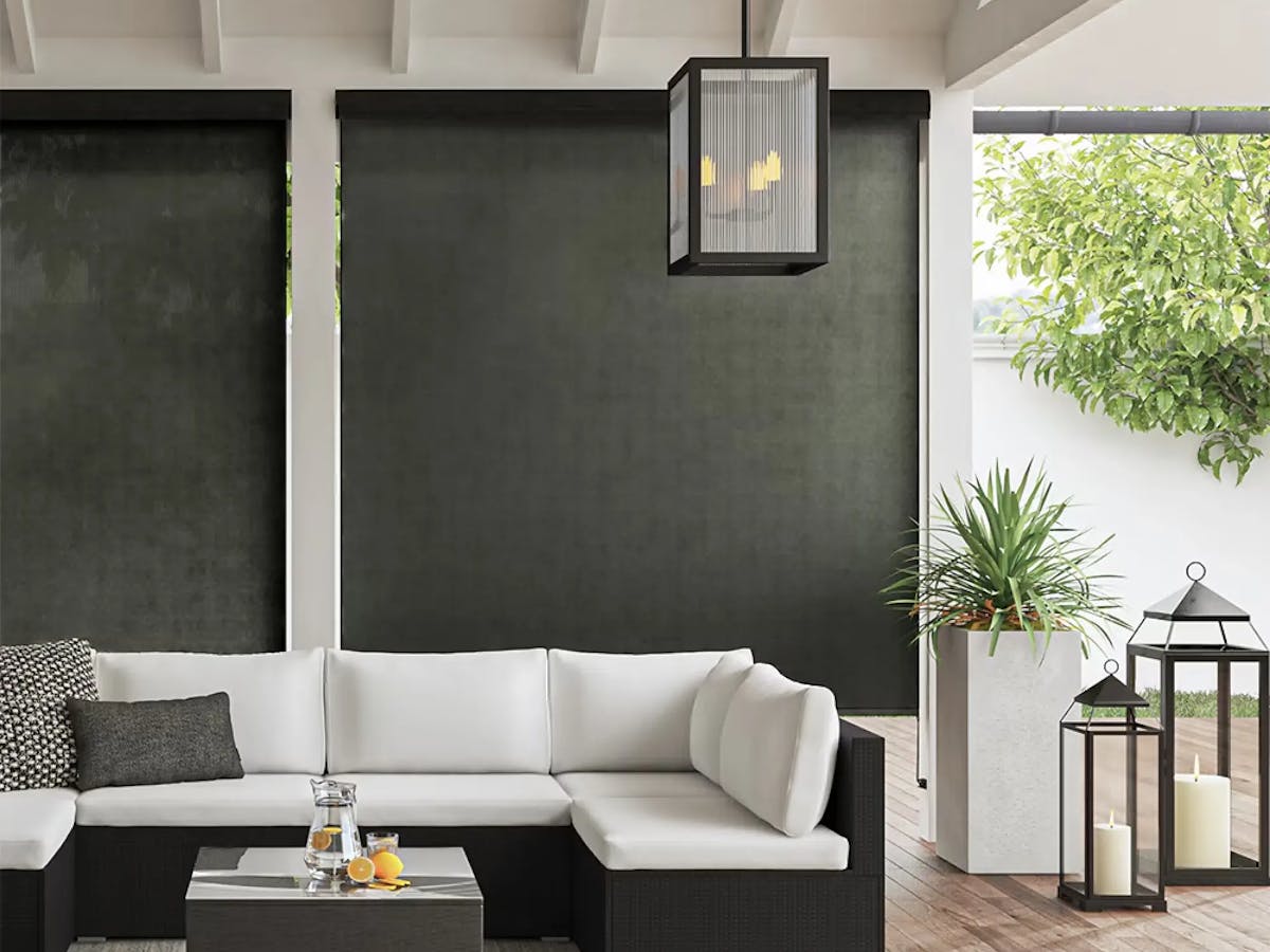 Outdoor patio with black and white furniture. There's a U-shaped couch with a glass-top coffee table in the center. Underneath the furniture is a gray woven rug. Behind the couch are white pillars with black outdoor shades.
