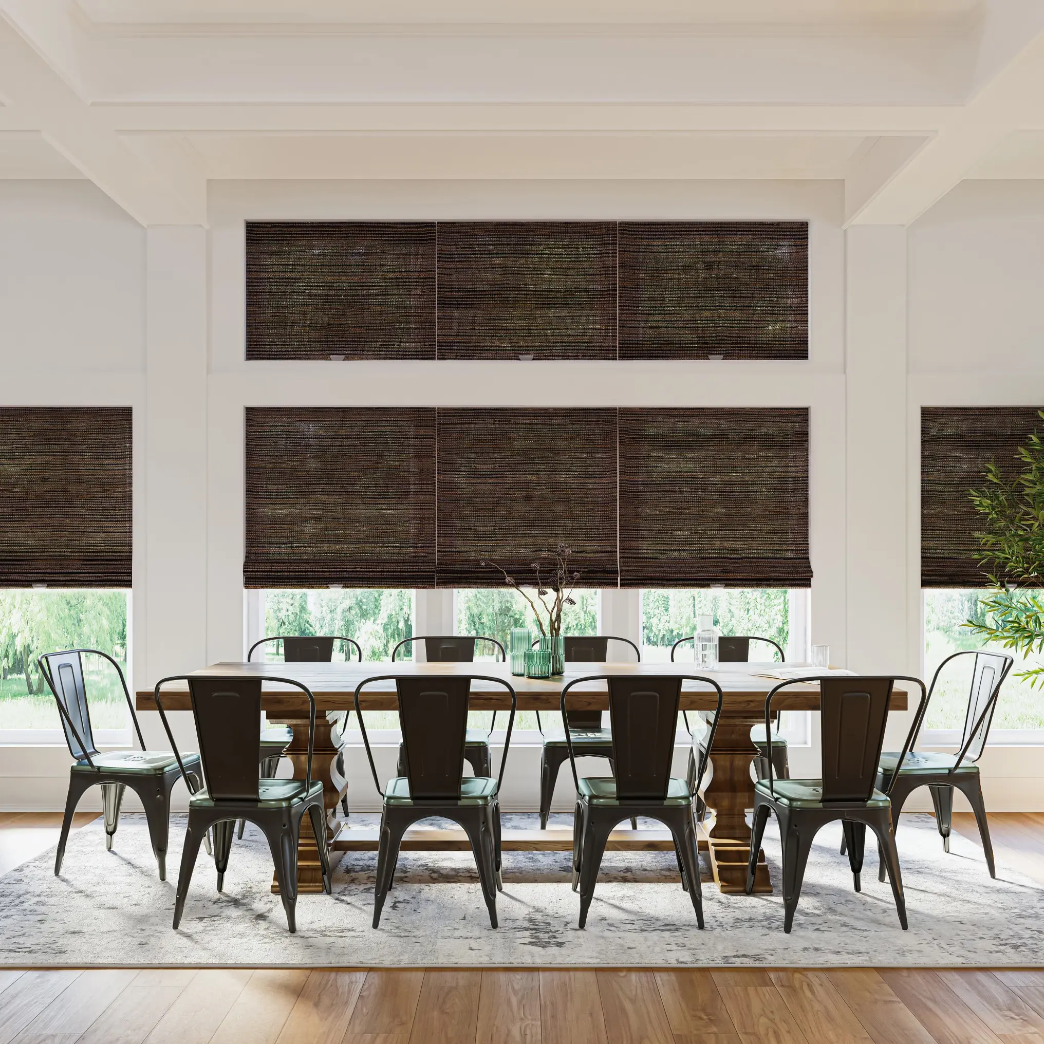 Dining room with a brown, 10-seater table surrounded by black metal dining chairs. Underneath the furniture is a patterned, gray rug. Behind the table is a wall of windows with dark brown woven wood shades.