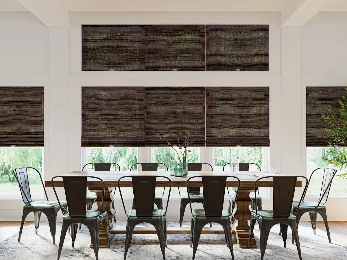 Dining room with a brown, 10-seater table surrounded by black metal dining chairs. Underneath the furniture is a patterned, gray rug. Behind the table is a wall of windows with dark brown woven wood shades.