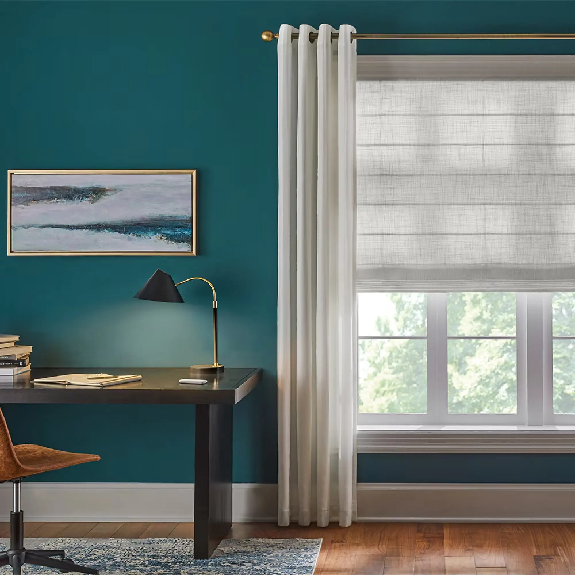 Office space with teal blue wall paint. On the left is a partial view of a dark colored desk. Above the desk is an abstract art piece. On the right is a window covered with gray roman shades layered behind white curtains.