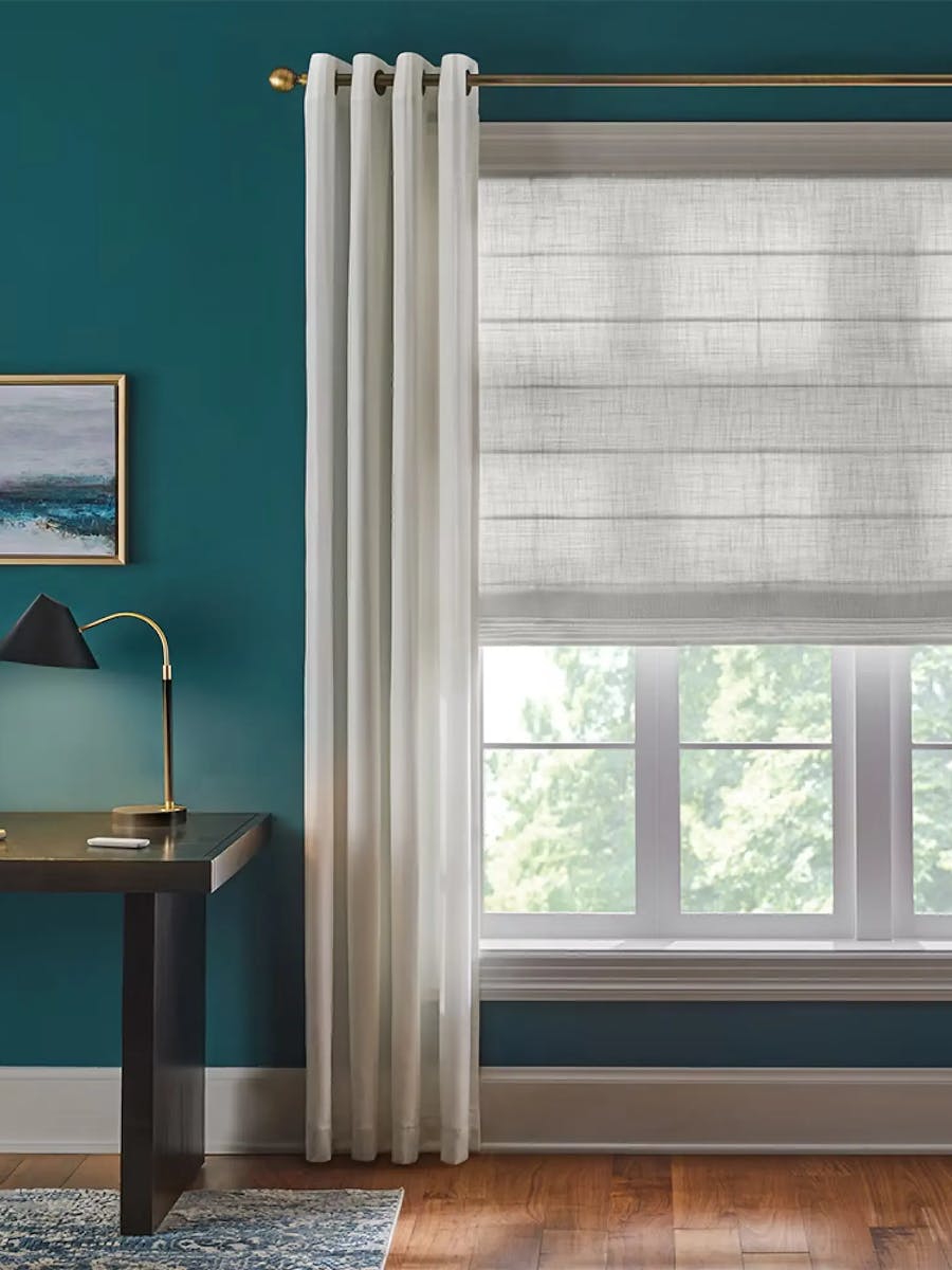 Office space with teal blue wall paint. On the left is a partial view of a dark colored desk. Above the desk is an abstract art piece. On the right is a window covered with gray roman shades layered behind white curtains.