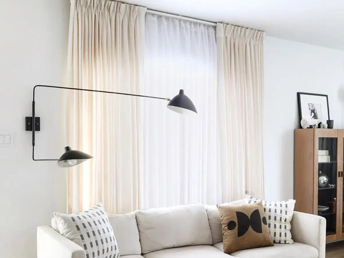 Bright living room featuring white curtains and double wall lamp hovering over a white couch with accent pillows.