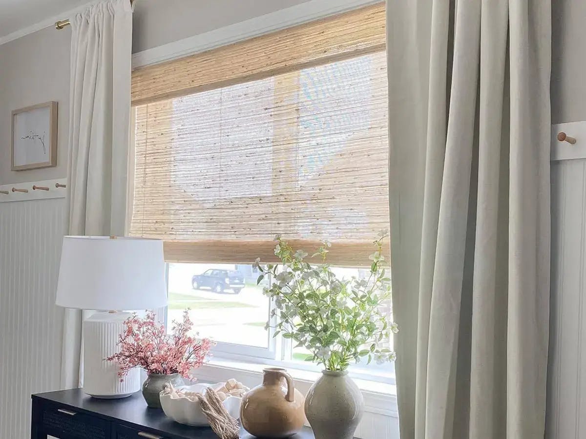 A window with woven wood shades, white curtains, and a table below featuring a lamp and flower vases.