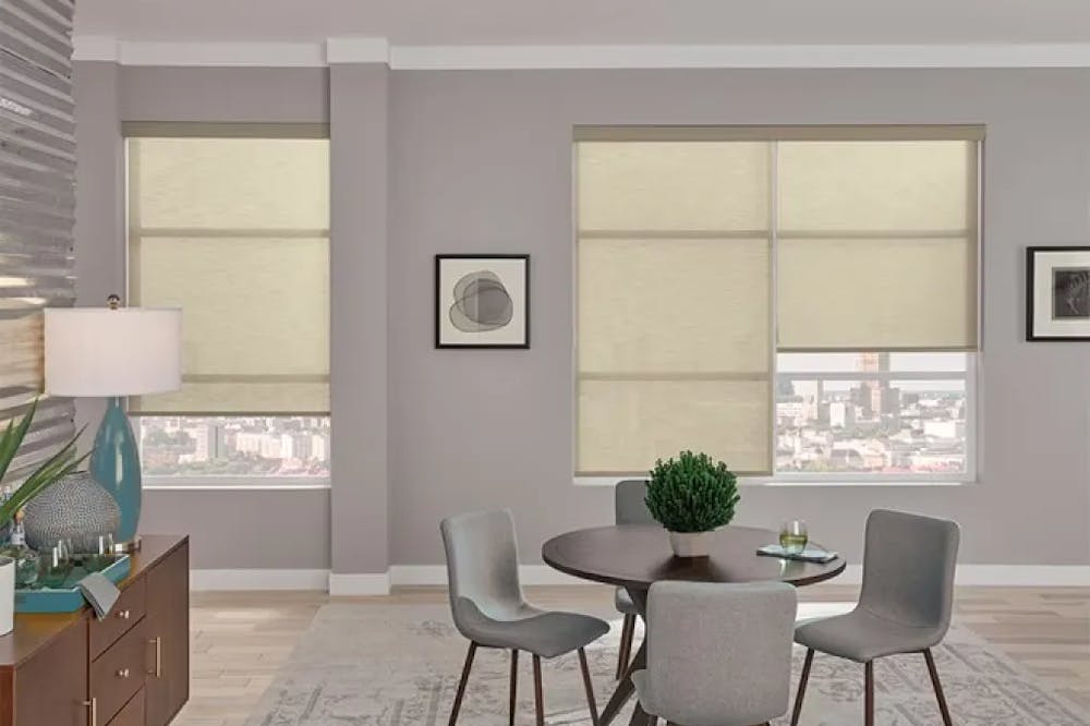 Dining room with mauve walls and cream roller shades on the windows. The dining table is dark wood with gray seats. There’s a patterned rug.
