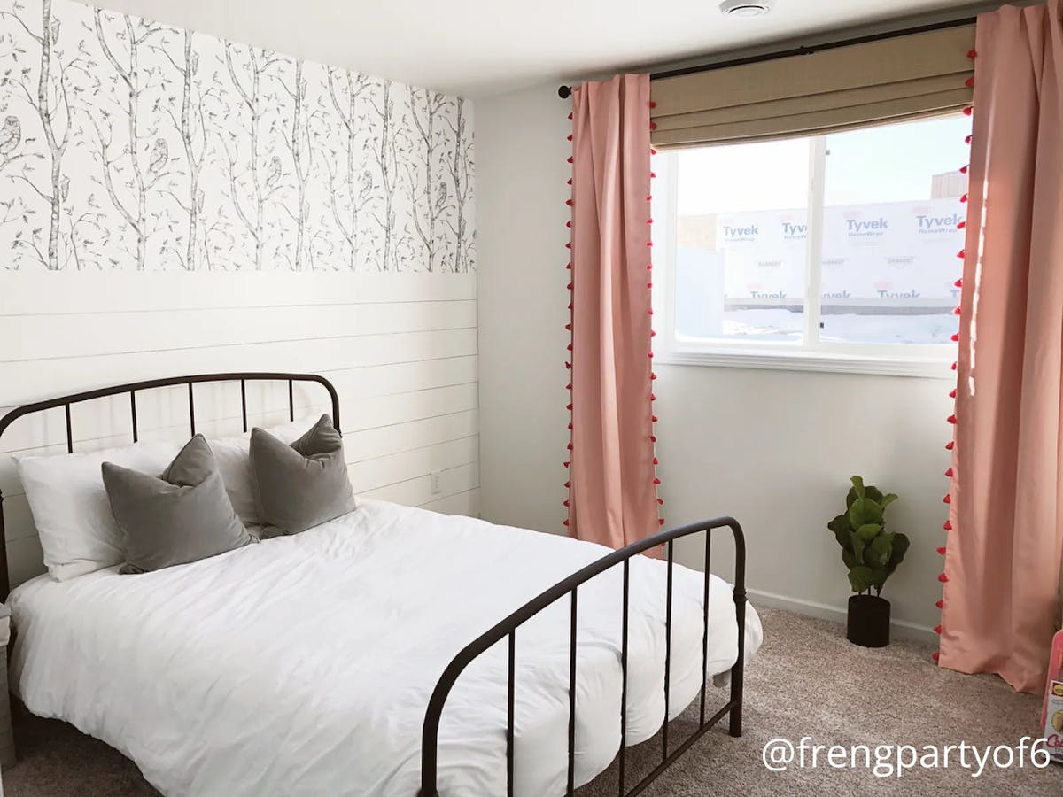 A bedroom with a pink carpet and wallpapered walls. The bed has white and gray bedding. There’s a window with raised woven wood shades and pink, tasseled floor-length curtains.