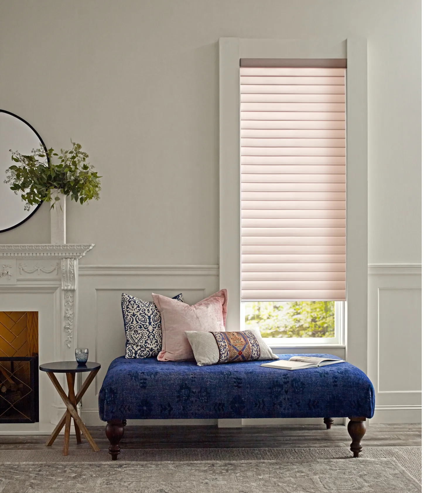 Window with light pink, sheer shades. In front of the window is a deep blue, cushioned bench with an assortment of pillows.
