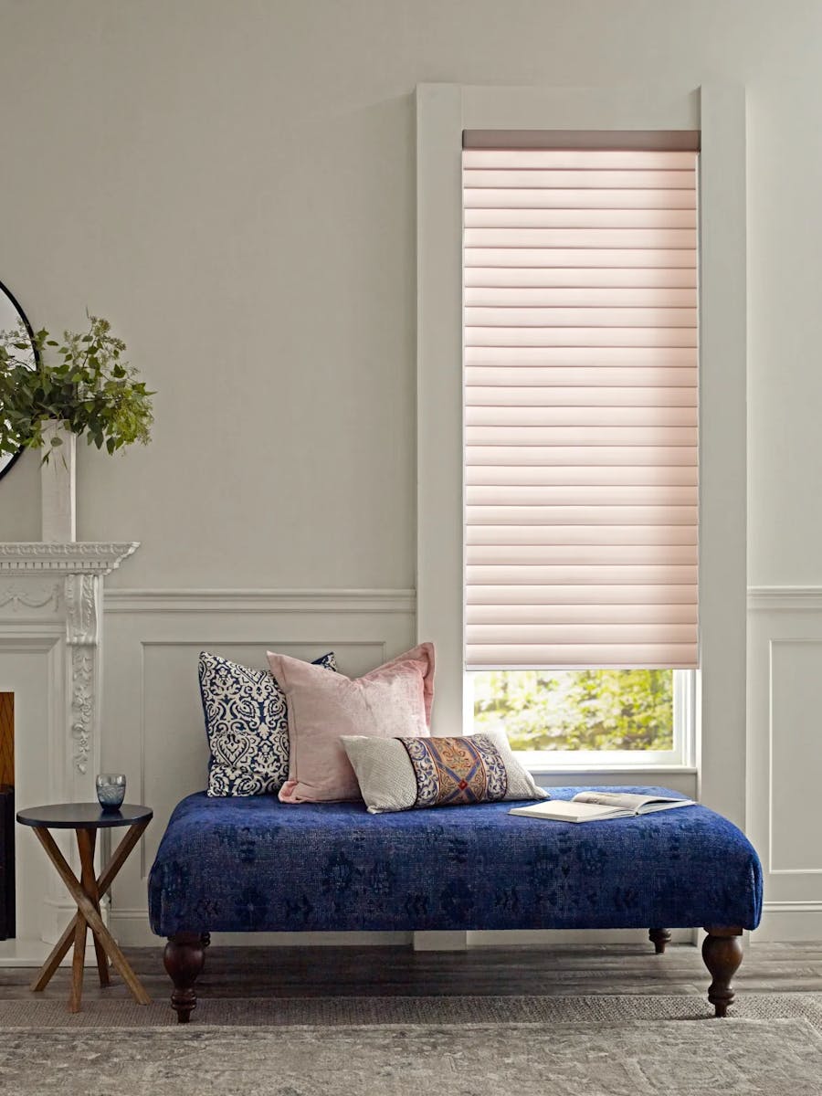 Window with light pink, sheer shades. In front of the window is a deep blue, cushioned bench with an assortment of pillows.