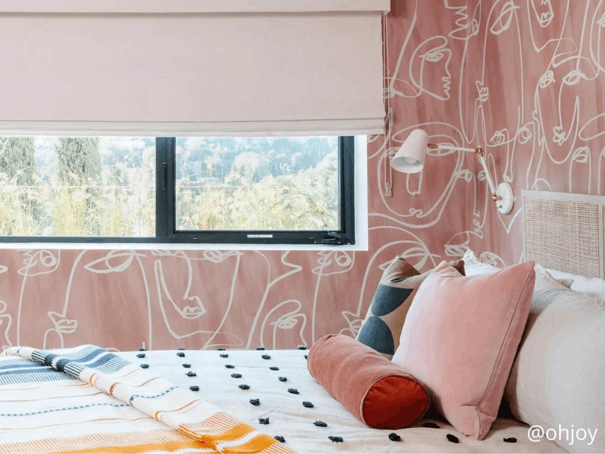 A bedroom with pink, patterned wallpaper. The window has a raised, cream-colored roman shade. The bed has a variety of patterned blankets and pillows.