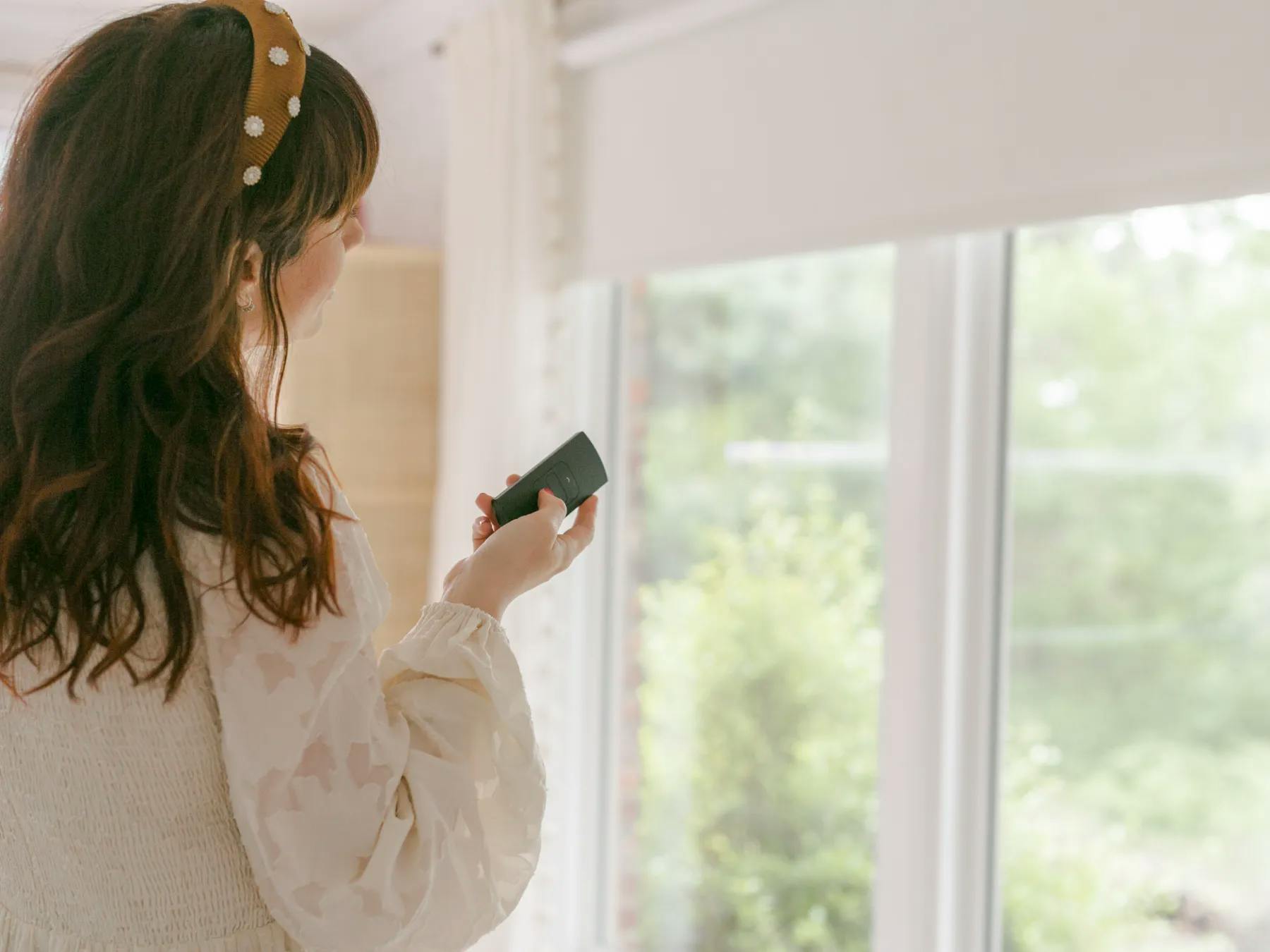 Woman with brown hair opening her white, motorized roller shades with a black remote.