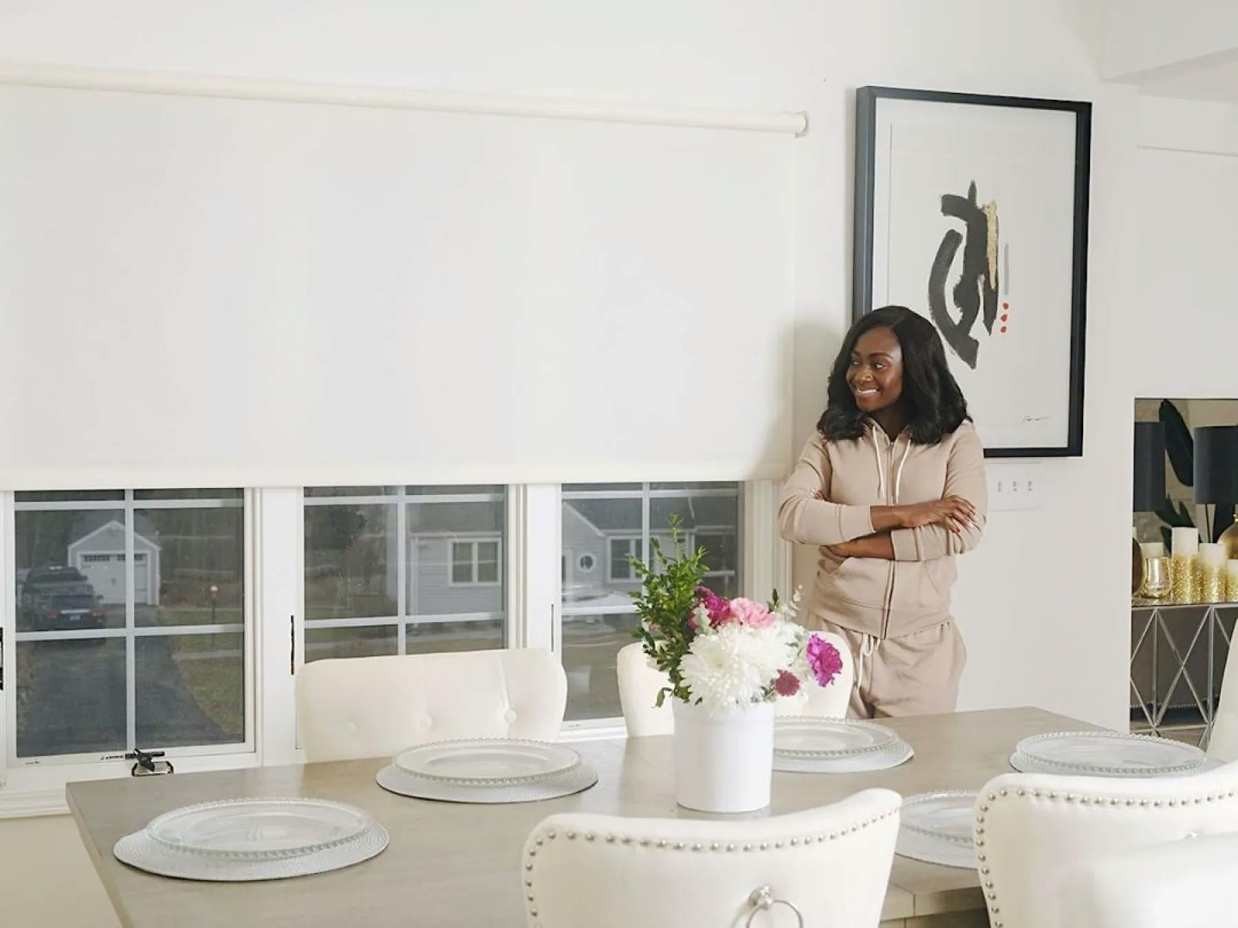A dining room with a greyish, brown table and white chairs. The table is set. A woman with dark skin is smiling, admiring her motorized white roller shades.