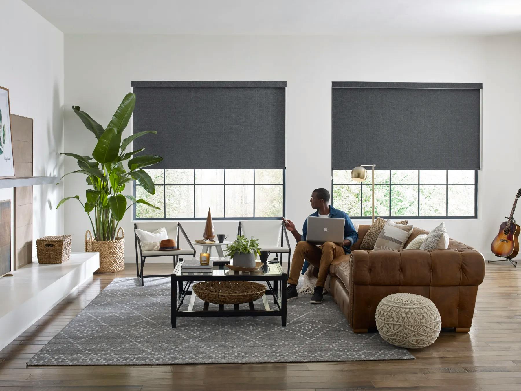 A dark skin man sits on his brown leather couch. He is using a remote to control his dark grey roller shades.