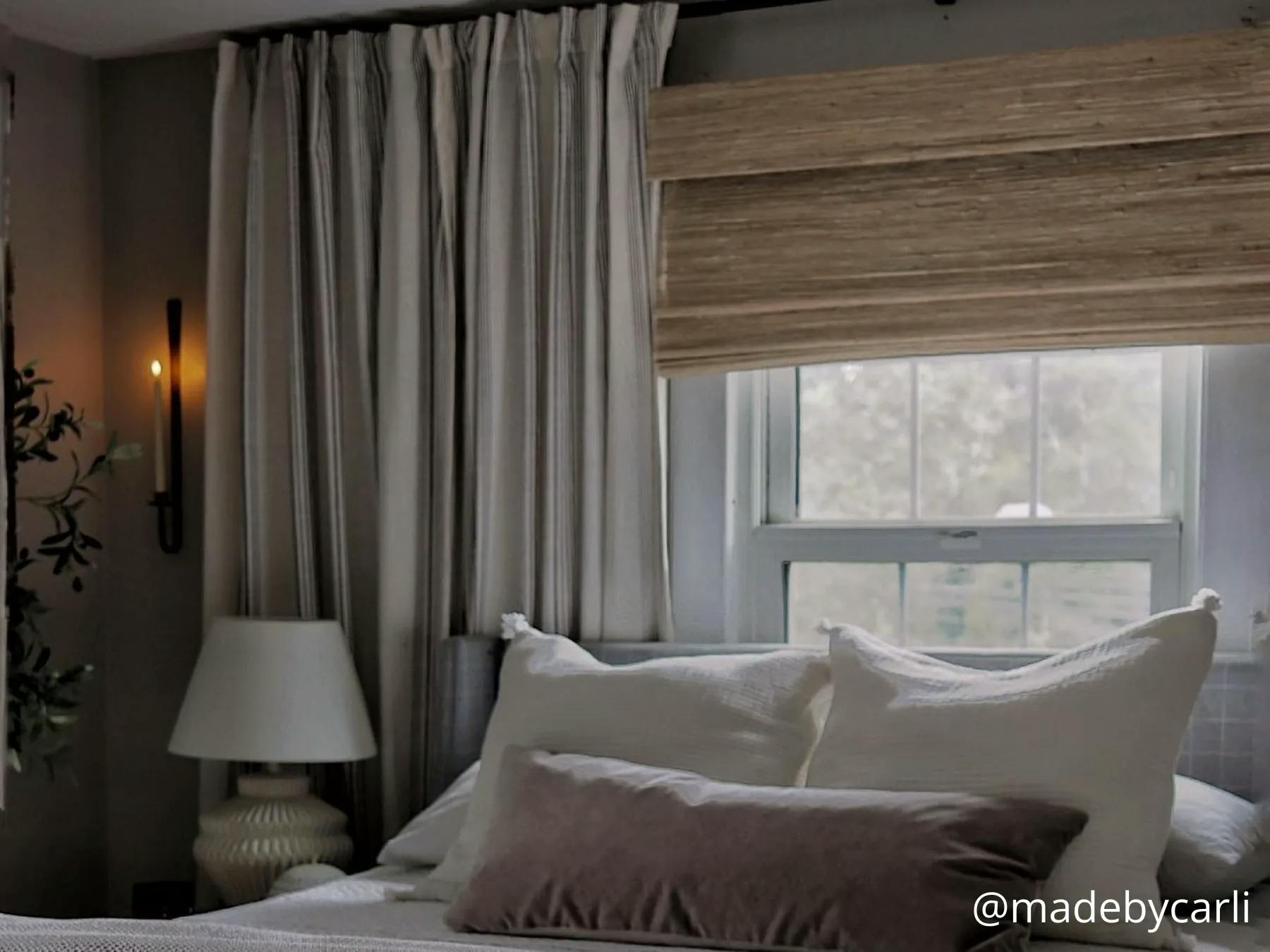 A bedroom with neutral bedding. The window is covered with a woven wood shade layered with neutral-colored drapes.