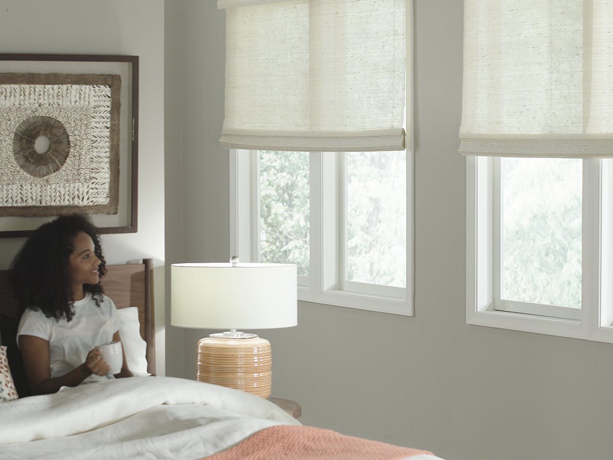 Woman in bed looking at motorized woven wood shades.