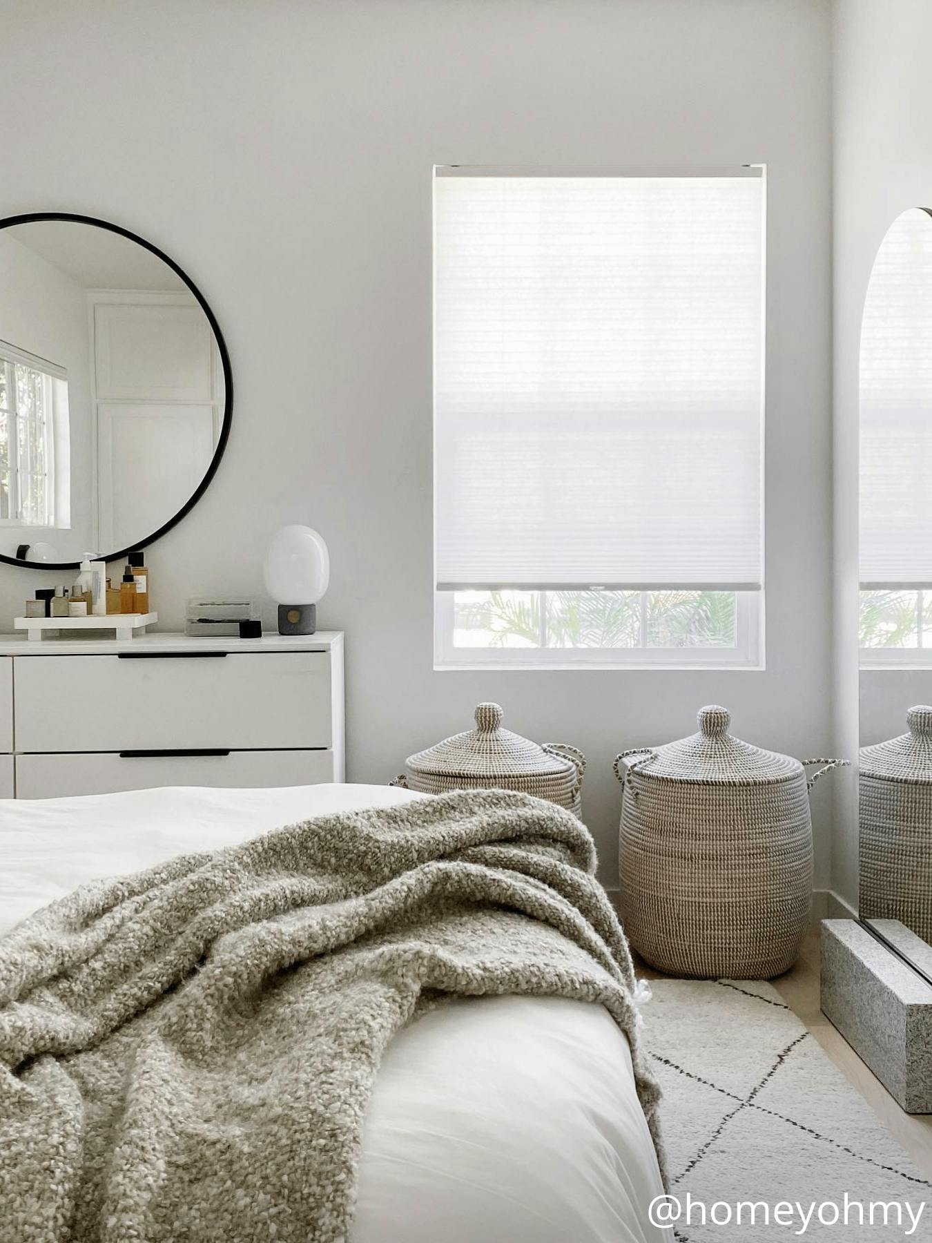A bedroom with white bedding and a white rug, featuring a round mirror above a white dresser and white, light-filtering cellular shades on the window.
