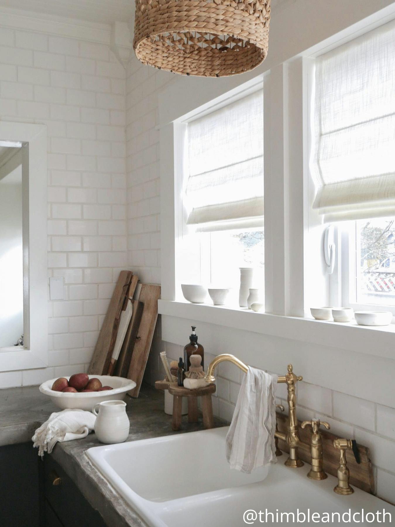 Kitchen counter with a white sink and gold hardware, surrounded by cutting boards and a fruit bowl. White roman shades cover the window.