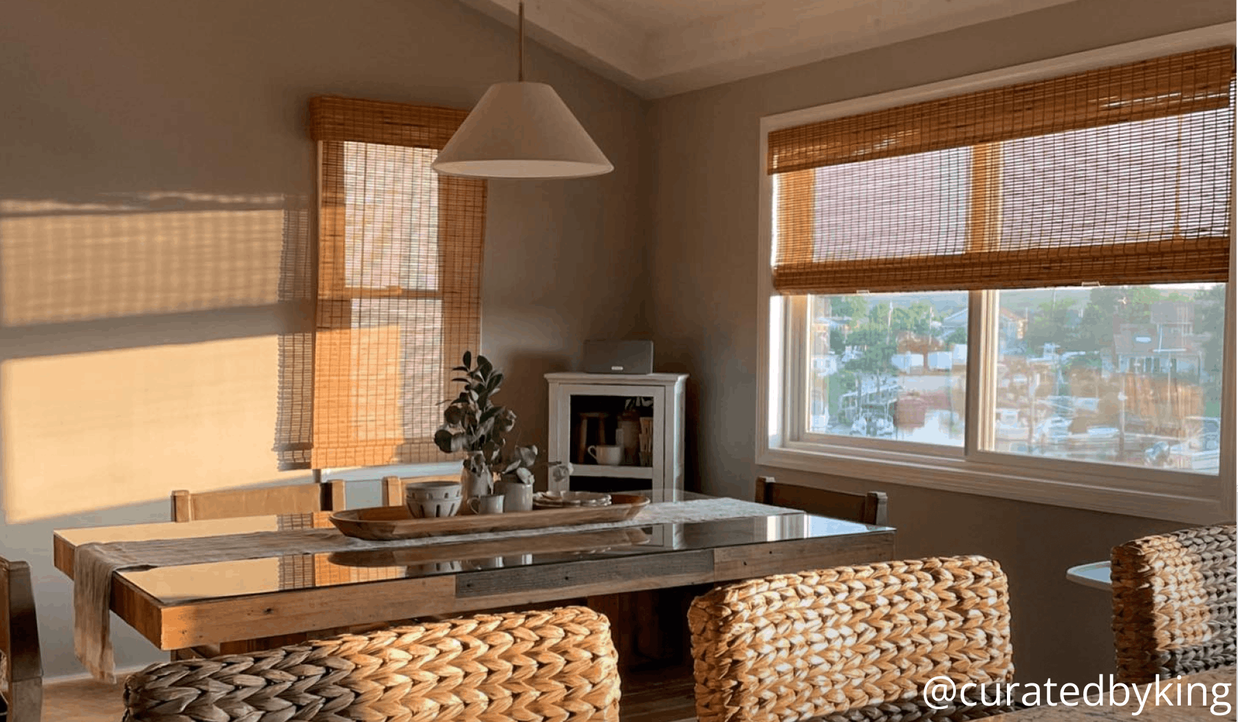 A dining room with a pendant light and warm golden hour light streaming through brown woven wood shades.