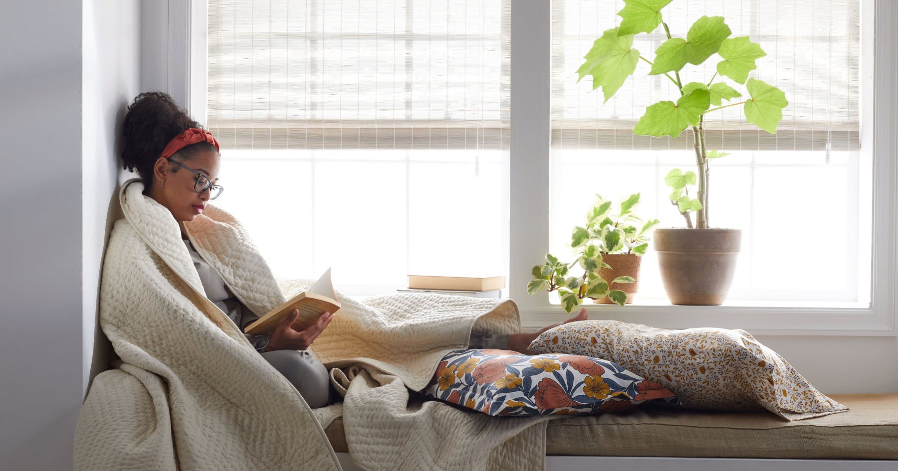 A woman wrapped in a blanket sits on a window seat, reading a book. Natural sunlight filters through woven wood shades, with leafy plants on the windowsill.