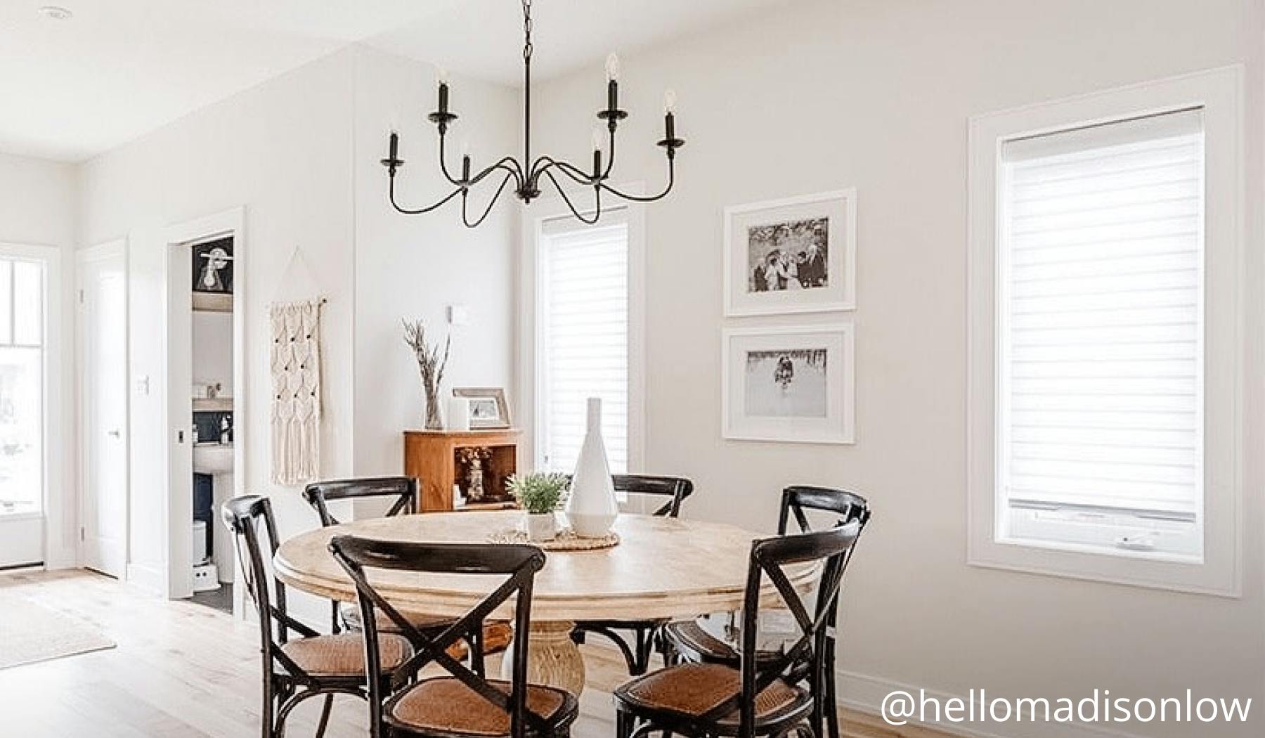 A dining room with a round table, six chairs and a candle chandelier overhead. The window has light-filtering shades.