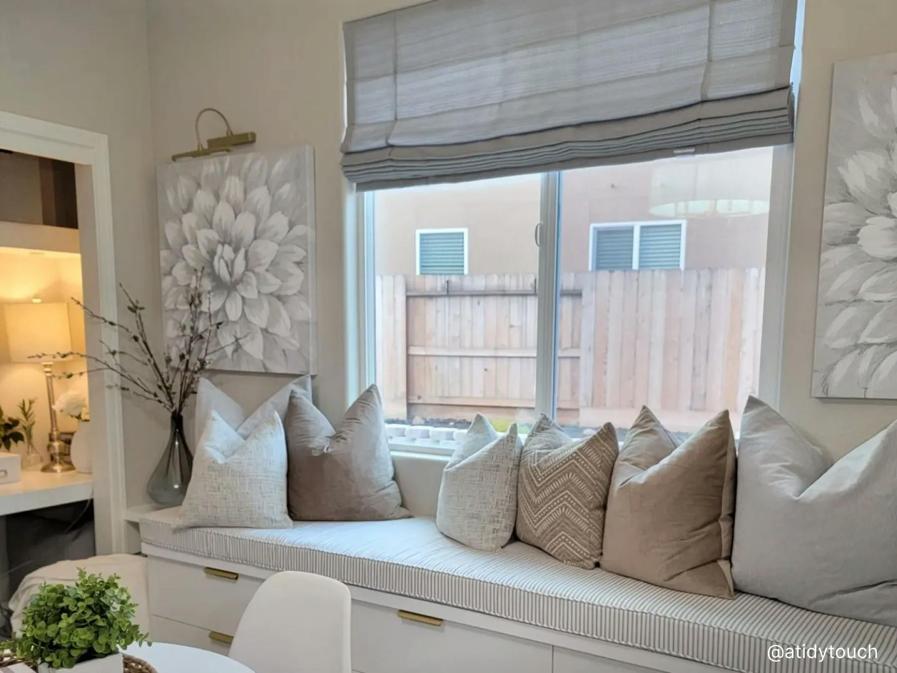 Cozy dining room with a white table, white chairs and window bench. The bench has neutral-colored pillows. Above the bench is a window with gray Roman shades. The window has flower paintings on both sides.