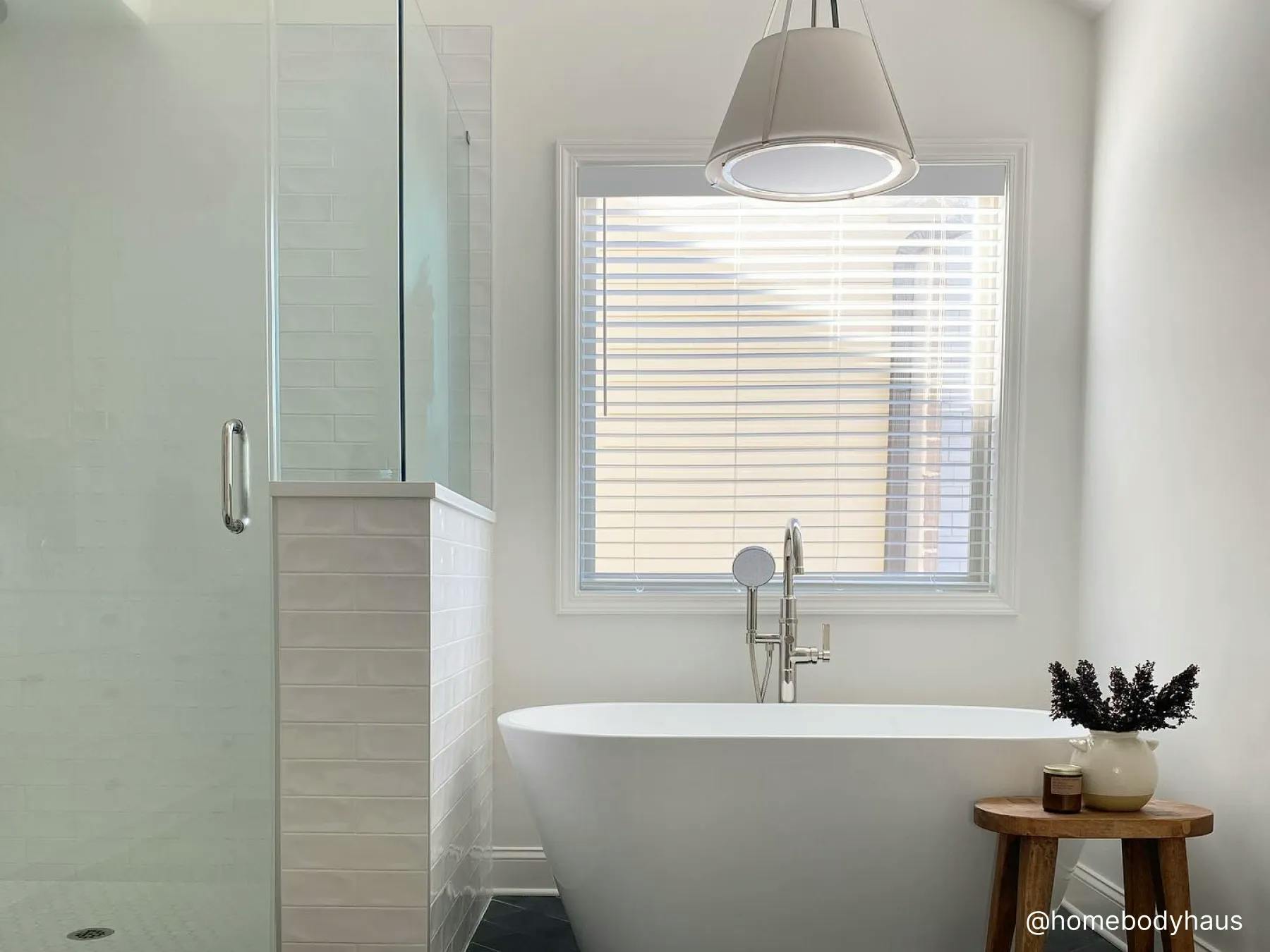 Minimalist bathroom with a white tub, glass shower and black tile floors. The window over the tub features white faux wood blinds. A white pendant light hangs over the tub.