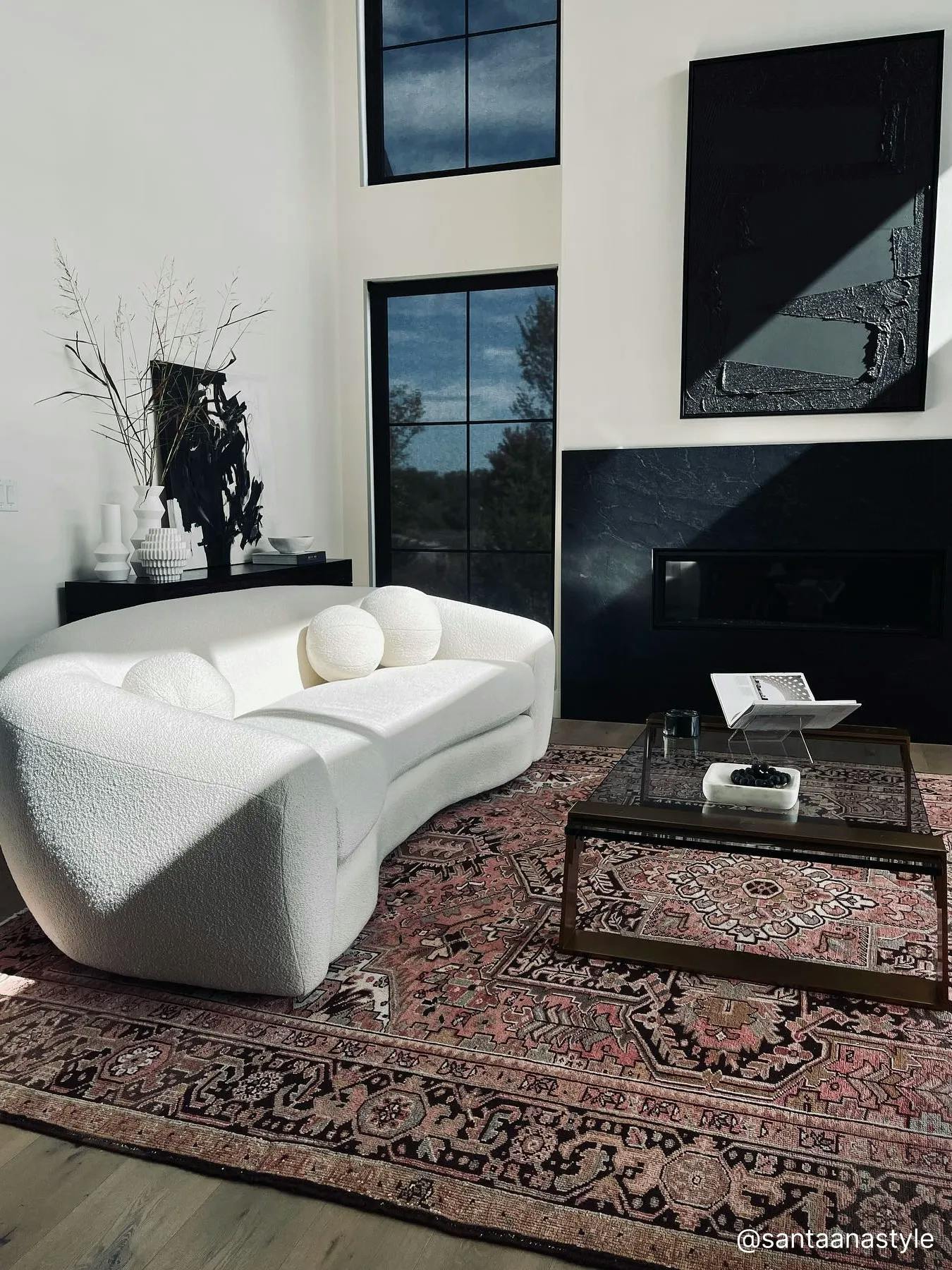 Modern living room featuring a white, rounded couch, a black, glass coffee table and a black, modern fireplace on the wall. The windows are covered with black, light-filtering solar shades.