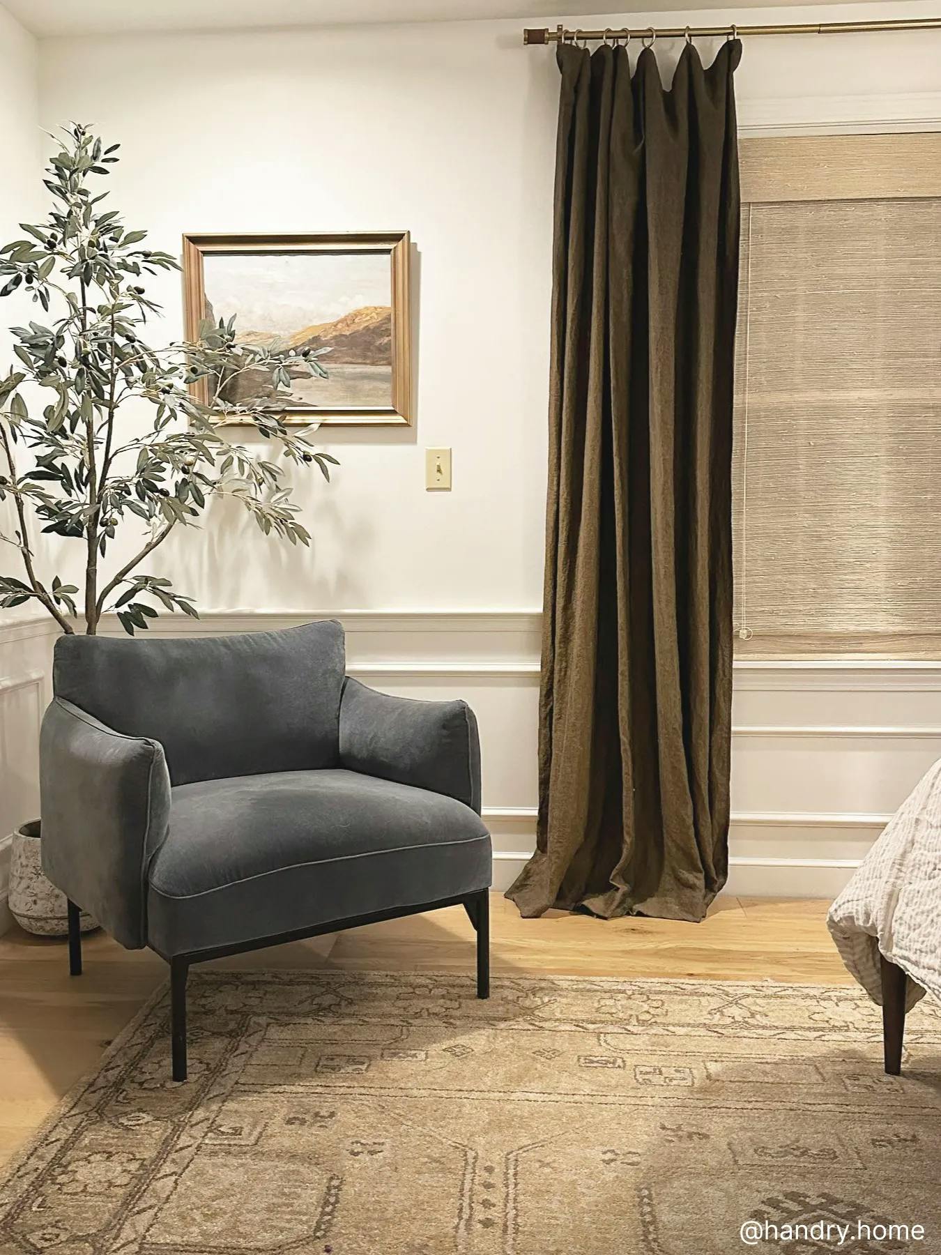 Bedroom with light wood floors, a patterned rug and gray reading chair. Behind the chair is a tall plant. The wall features art and a window covered with dark brown curtains layered over light woven wood shades.