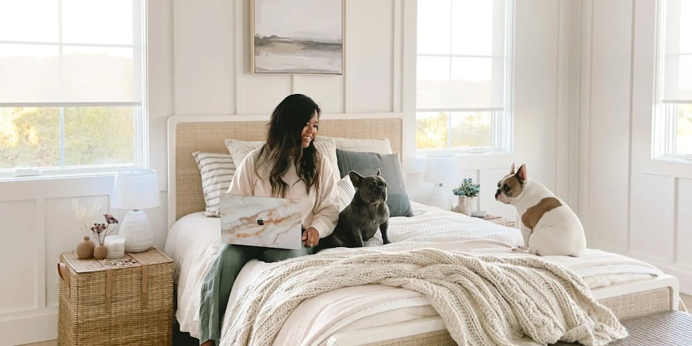 A woman and her dogs sitting on a bed with cream bedding. The room is bright and has windows with white, light filtering roller shades.