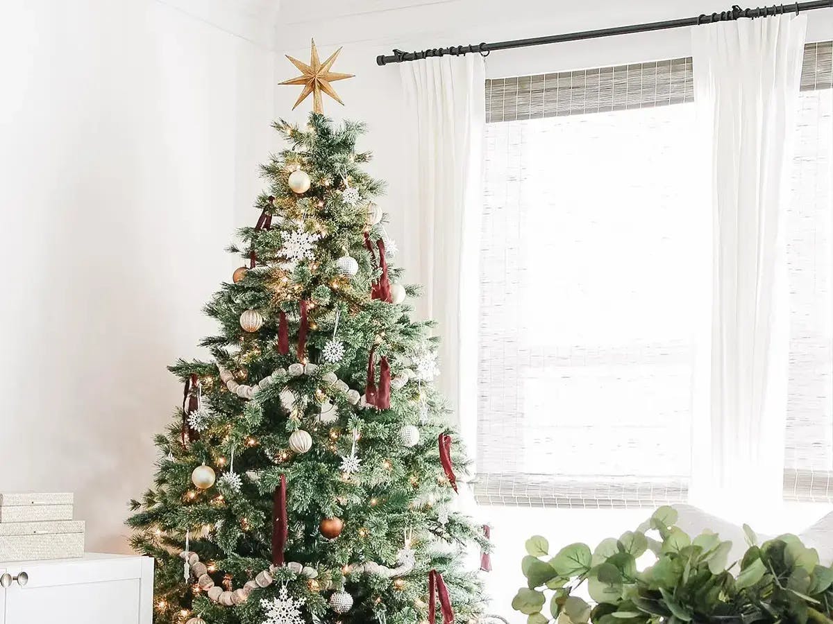 A decorated Christmas tree with a golden star topper, white snowflake ornaments, red ribbons, and soft white lights stands in a bright, minimalist living room.