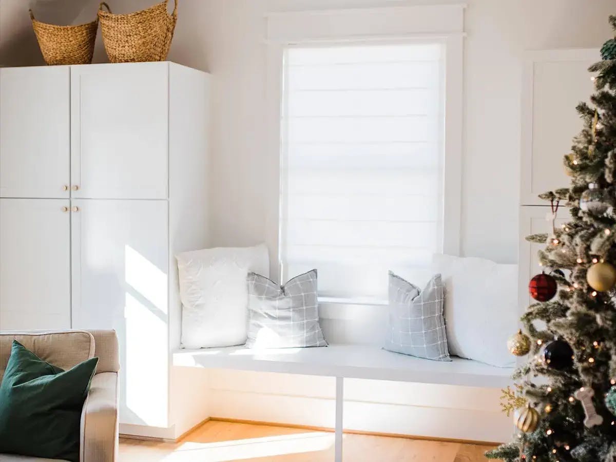 A minimalist space with a white bench by a window, with solid pillows, storage cabinets, and a decorated Christmas tree.