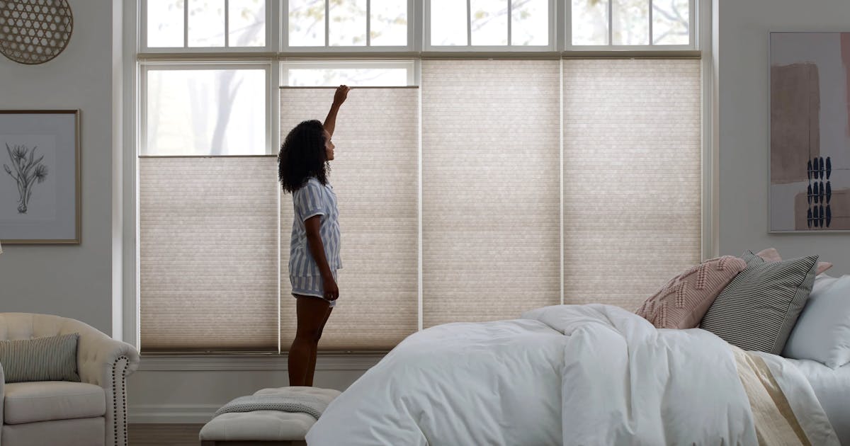 A woman adjusting her cellular shades in her bedroom. The bedroom has a neutral-colored bed, wooden nightstand, chair, lamp, wall decorations and cellular shades on four windows.