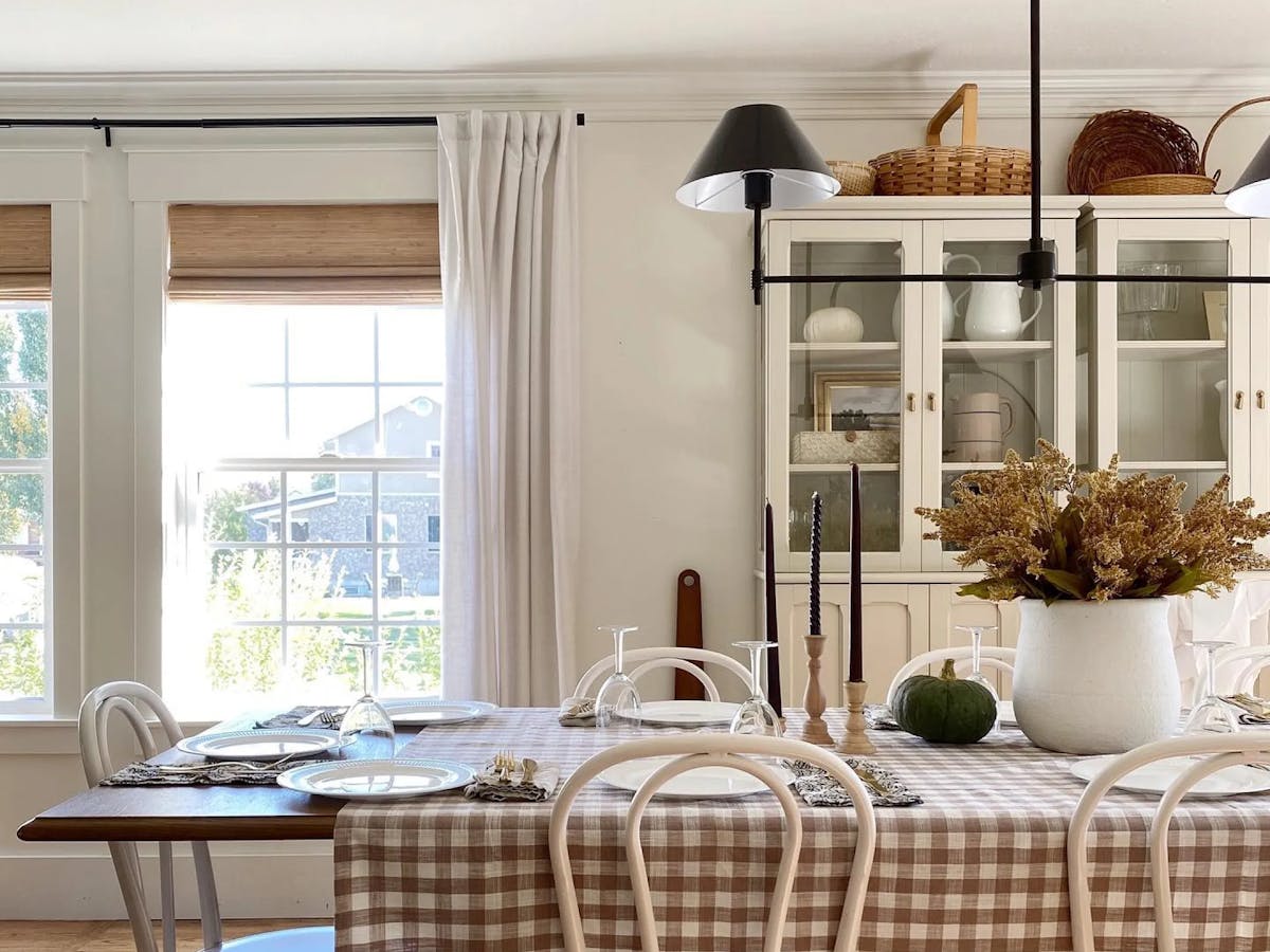 Dining room with a white China cabinet filled with white table wear. The dining table features a gingham tablecloth, autumnal centerpiece and candles. The window features open woven wood shades with white curtains.)