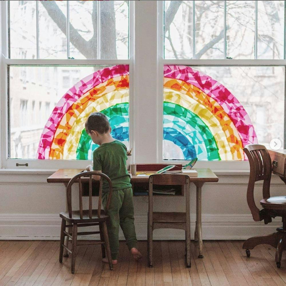 young child tapes colored pieces of tissue paper to a window in the shape of a rainbow.