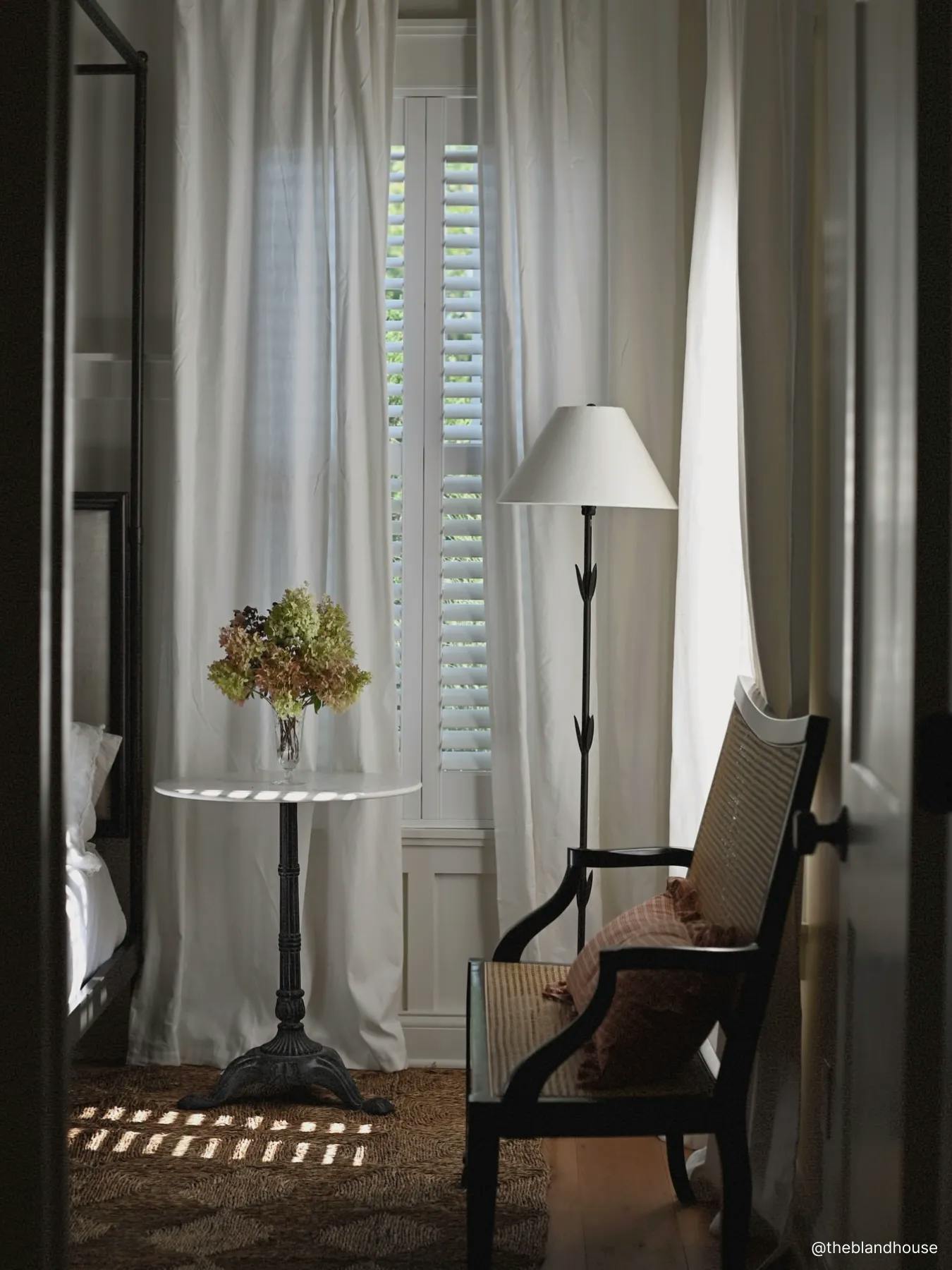 Window with faux wood blinds layered beneath white sheer shades. In front of the window is a side table with a vase of flowers on it as well as a lamp.