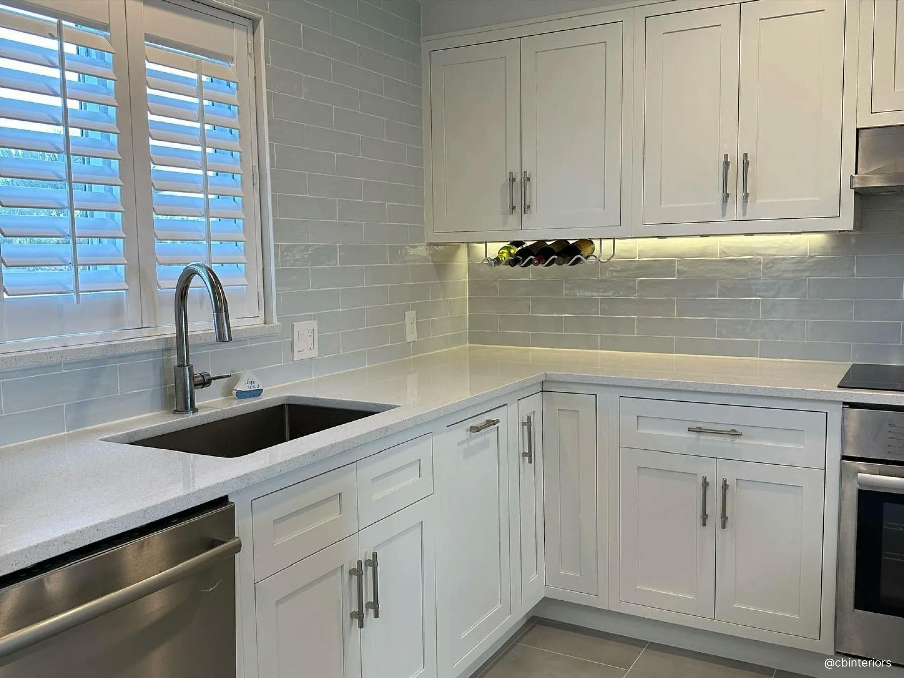 Kitchen with light gray, marble countertops and white cabinets. Above the stainless steel sink is a window with white interior shutters.