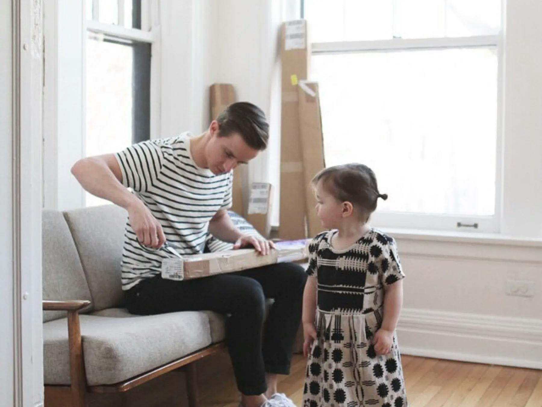 A man sitting in front of two uncovered windows, opening a box of blinds. Standing in front of him is his young daughter.