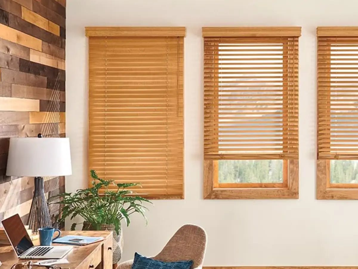 Office space with light brown wooden blinds. There's a desk with a laptop, mug and papers. The chair in front of the desk has a blue pillow.