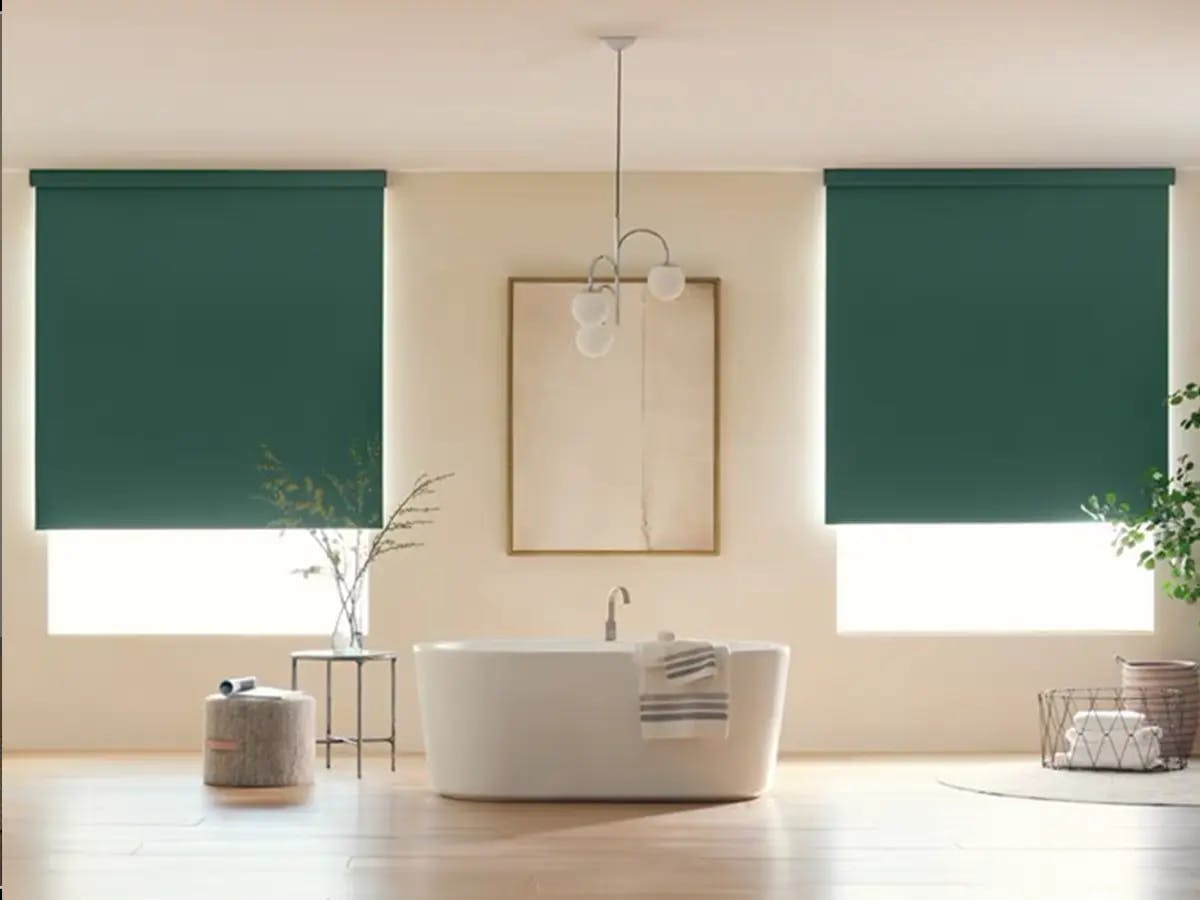 Bathroom with a large white tub. Above the tub is a modern abstract art piece. To the left and right of the art piece are large windows covered with emerald green roller shades.