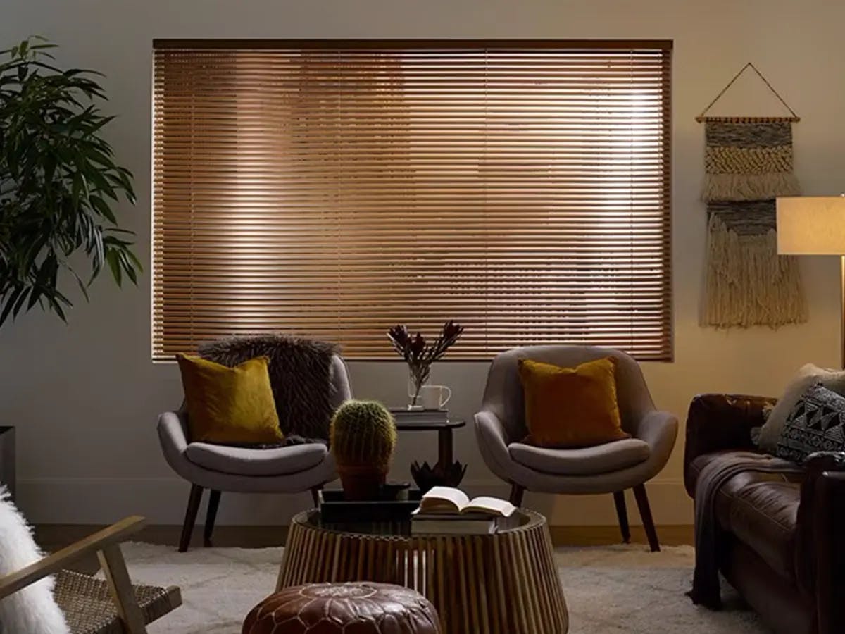 Dark brown faux wood blinds covering a large window. Below the window are two gray armchairs with neutral-colored pillows.