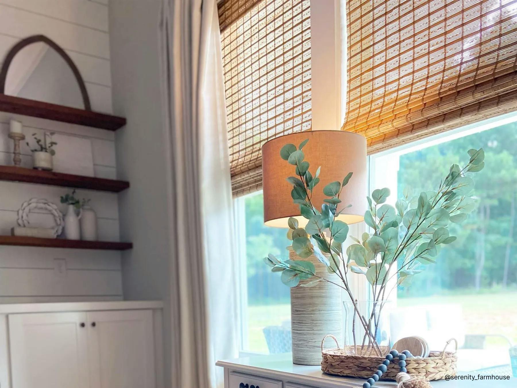 A window with woven wood shades. In front of the window is a table with a lamp and glass vase with eucalyptus leaves.
