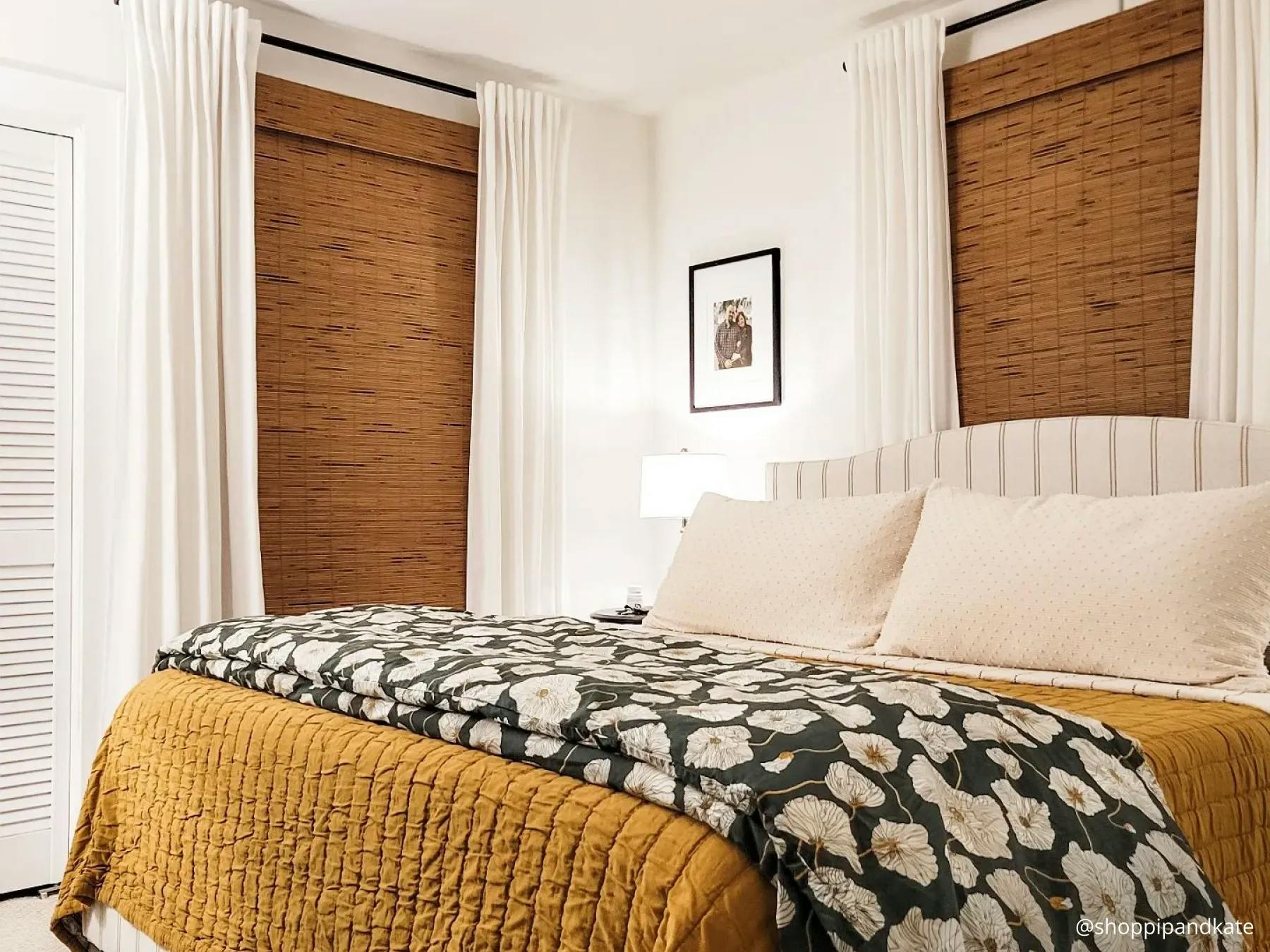 Bedroom with white, cream and warm-tone sheets. There's a floral throw blanket across the foot of the bed. The windows have  white curtains layered over blackout woven wood shades.