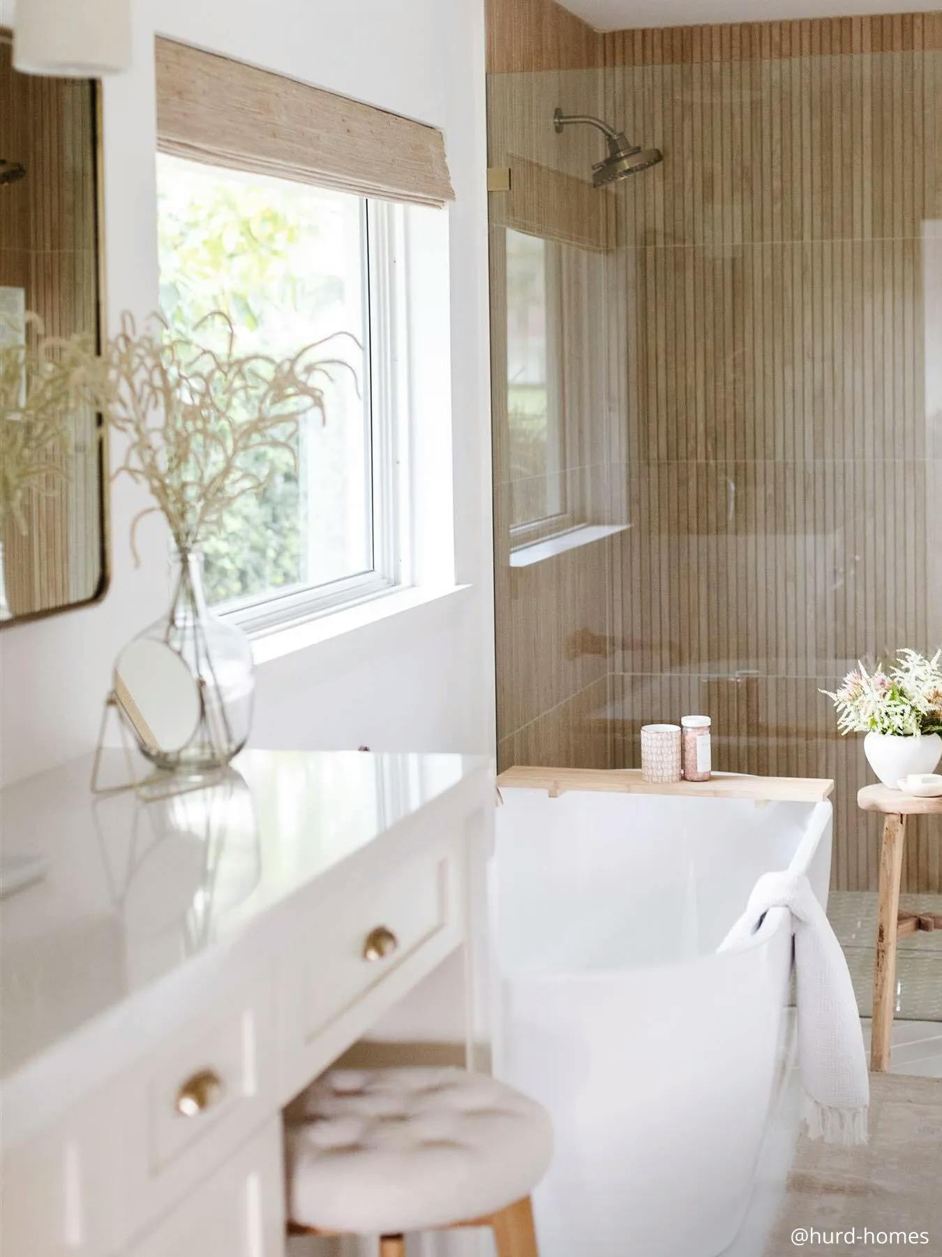 A neutral colored bathroom with a soaking tub and glass walk-in shower. Above the top is a window with raised woven wood shades. 