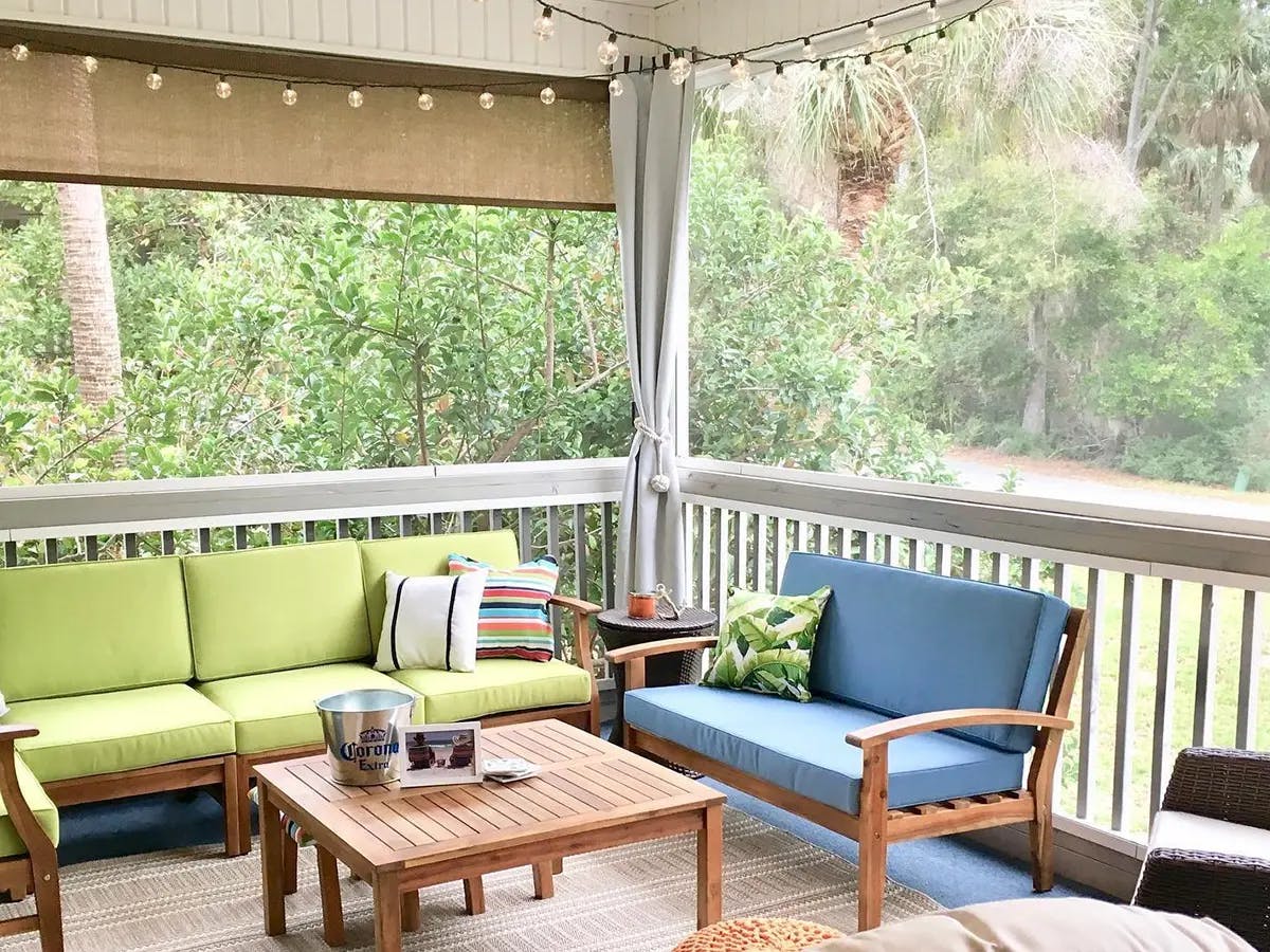 A porch featuring colorful cushioned seating, a wooden coffee table, string lights and greenery outside.