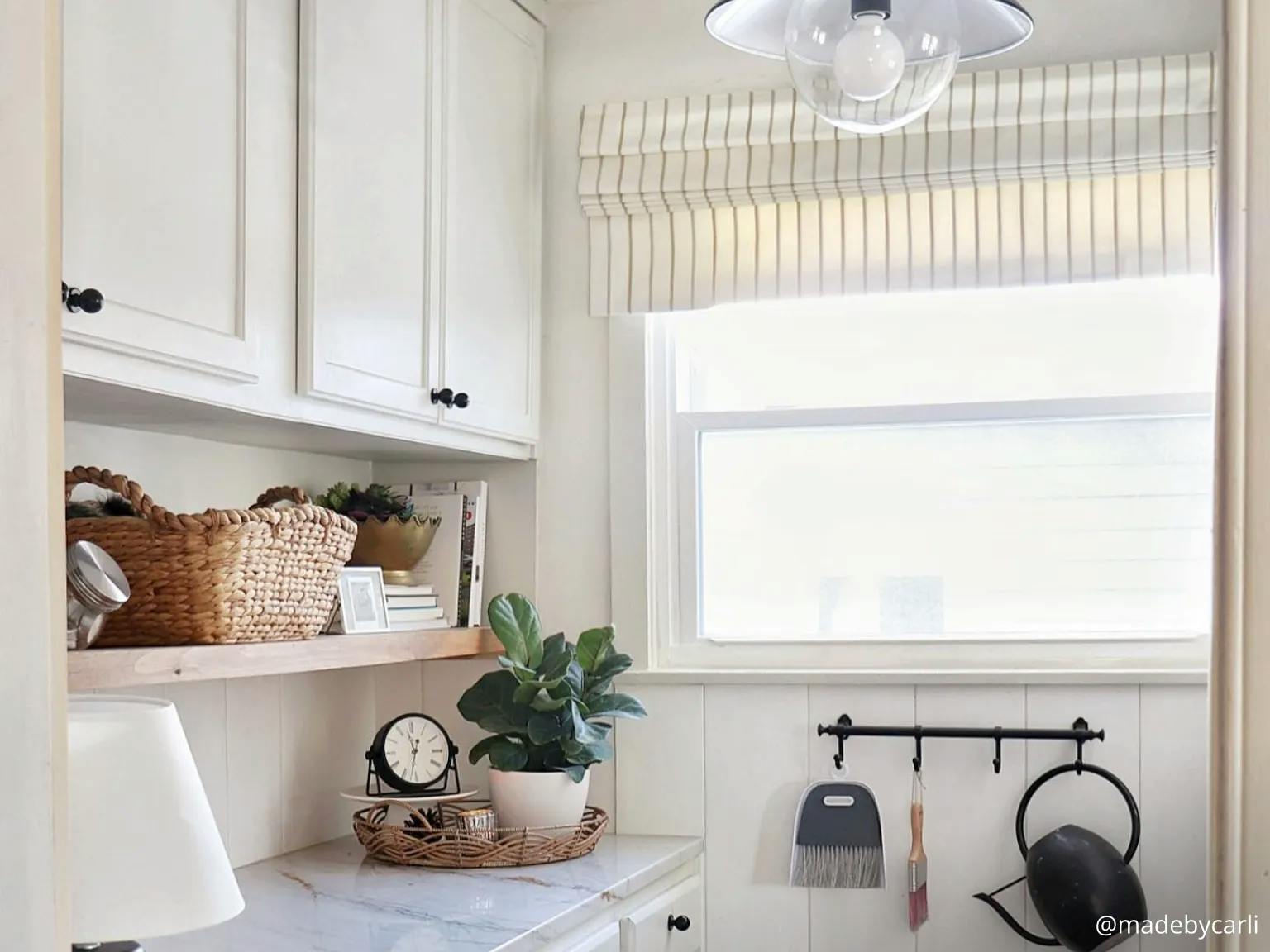 Laundry room with marble countertops, white shiplap walls and white cabinets with black hardware. The various decorations feature wood, wicker and neutral colors. The window has a striped roman shade in white.